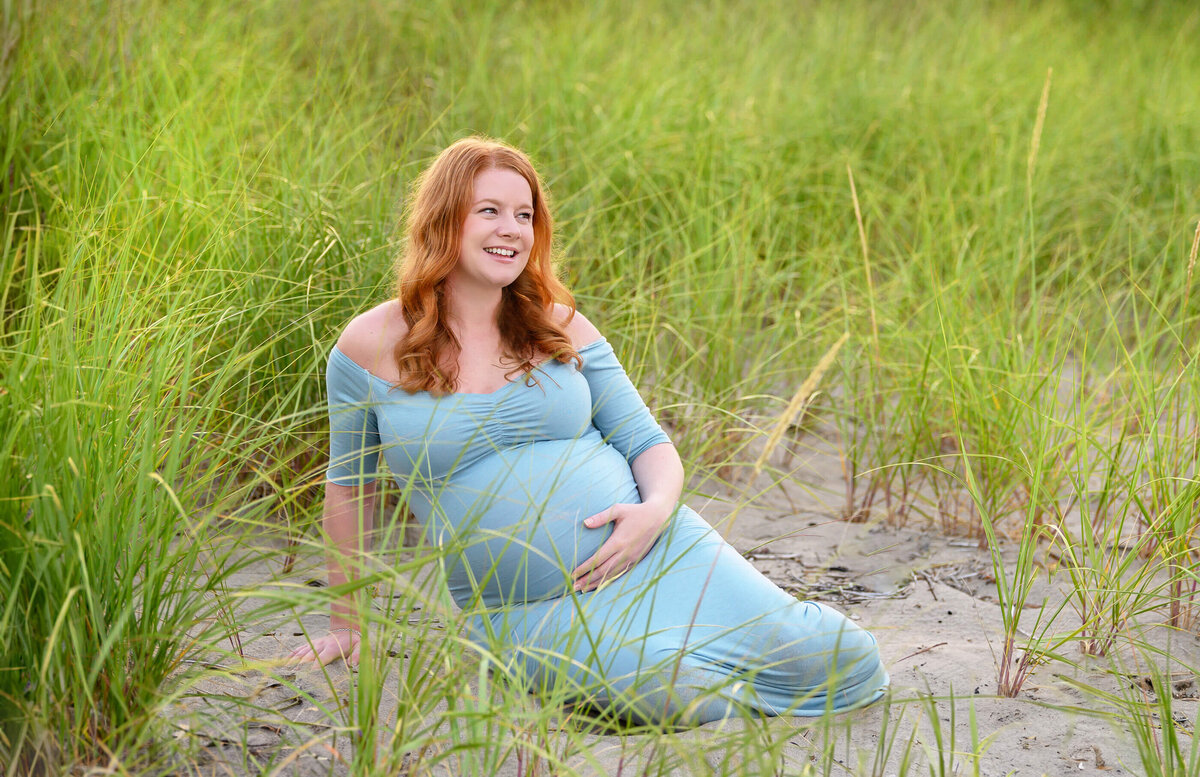 pregnant woman with red hair wearing a blue dress sitting in the sand with tall grass behind her for outdoor beach maternity photography session in Burlington