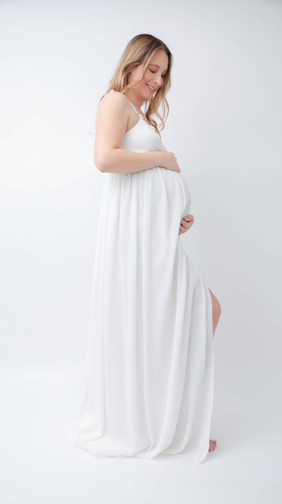 Pregnant mom wearing a white flowing dress and posed with one hand under belly and one hand above belly.