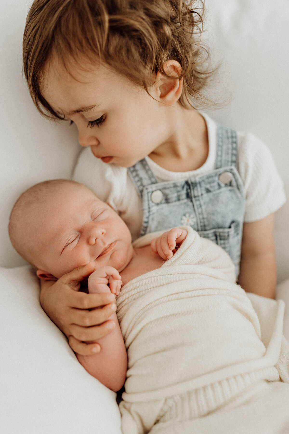A little boy holds his newborn baby brother wearing cute denim overalls while lucy jane takes candid newborn photographs of them in canterbury 