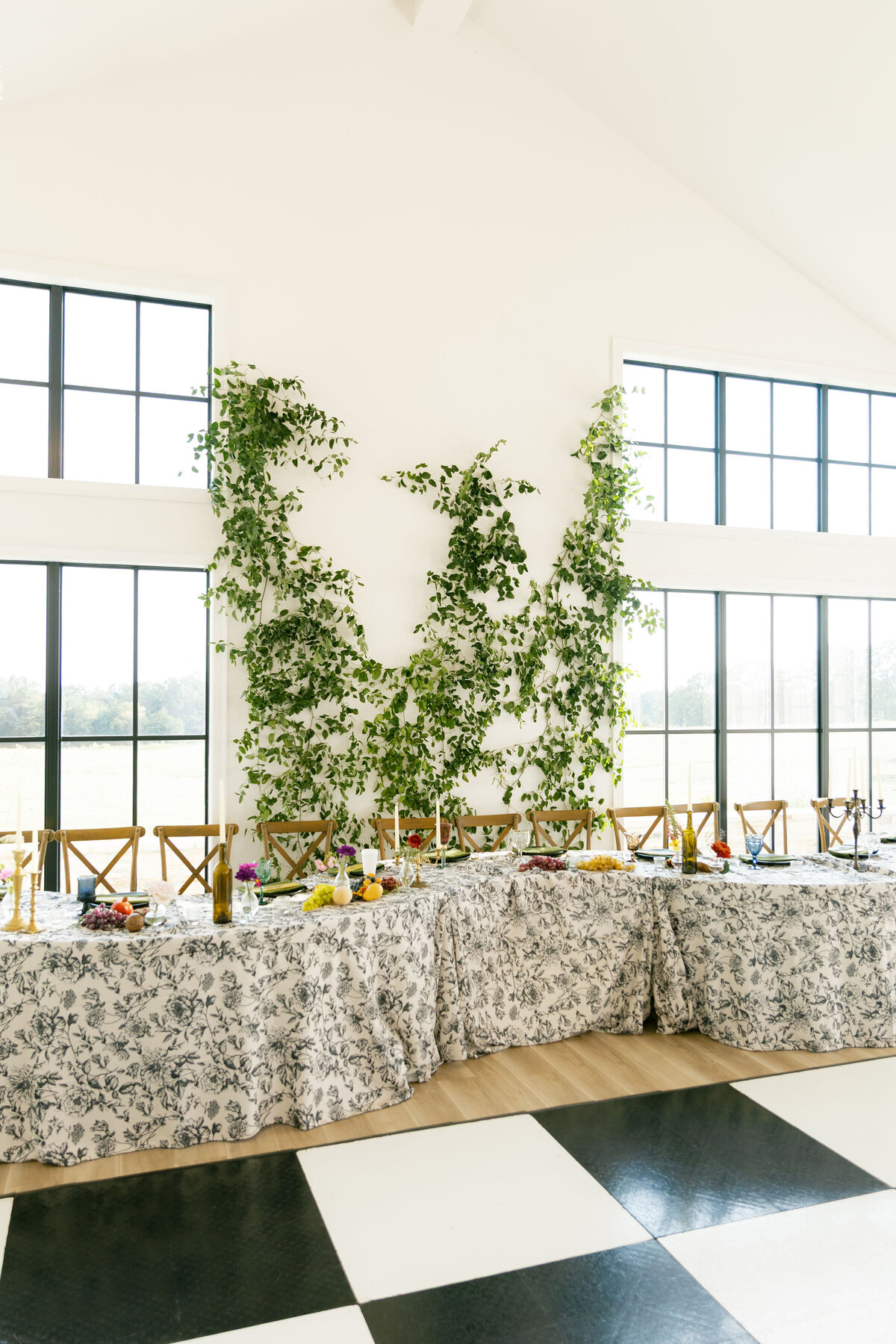 Reception head table decorated with toile linens, colorful fruit-inspired centerpieces, taper candles, and a lush greenery installation climbing the white wall in a bright modern venue.