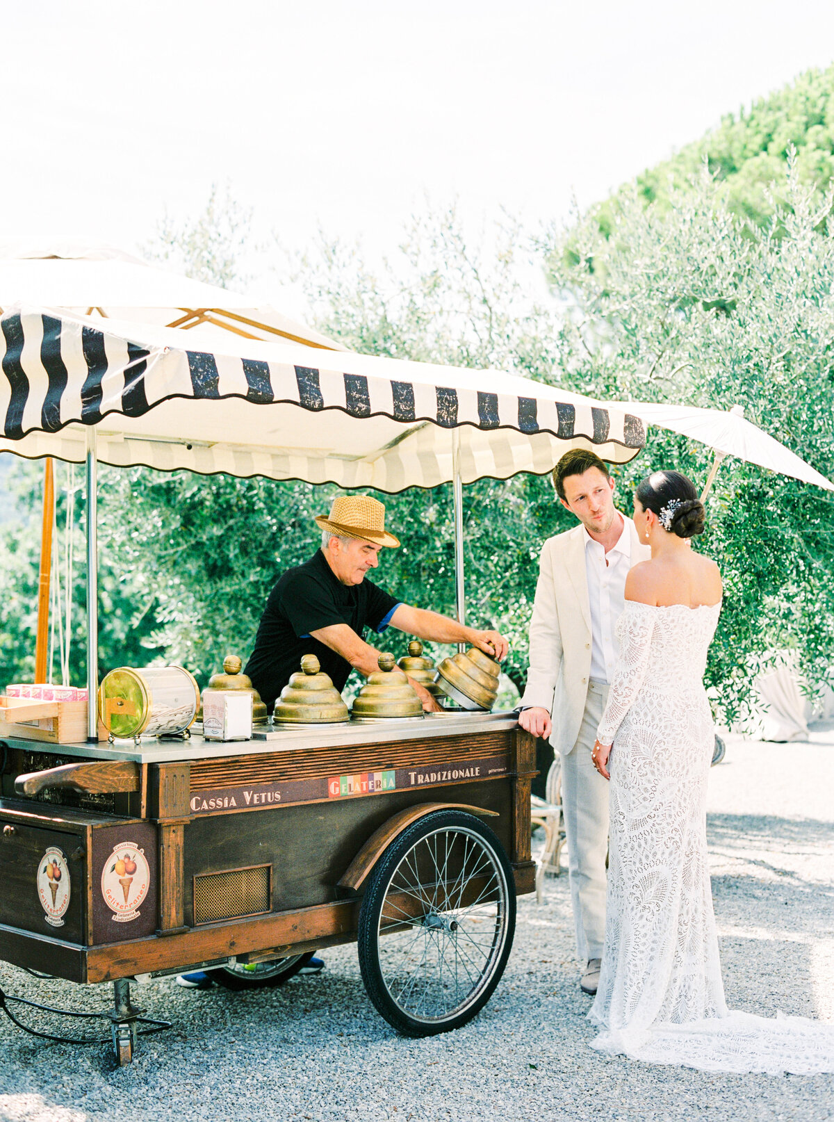 Film photograph of Bride in all lace wedding dress and parasol with groom in lite tan wedding suit ordering from a gelato truck at their wedding photographed by Italy wedding photographer at Villa Montanare Tuscany wedding
