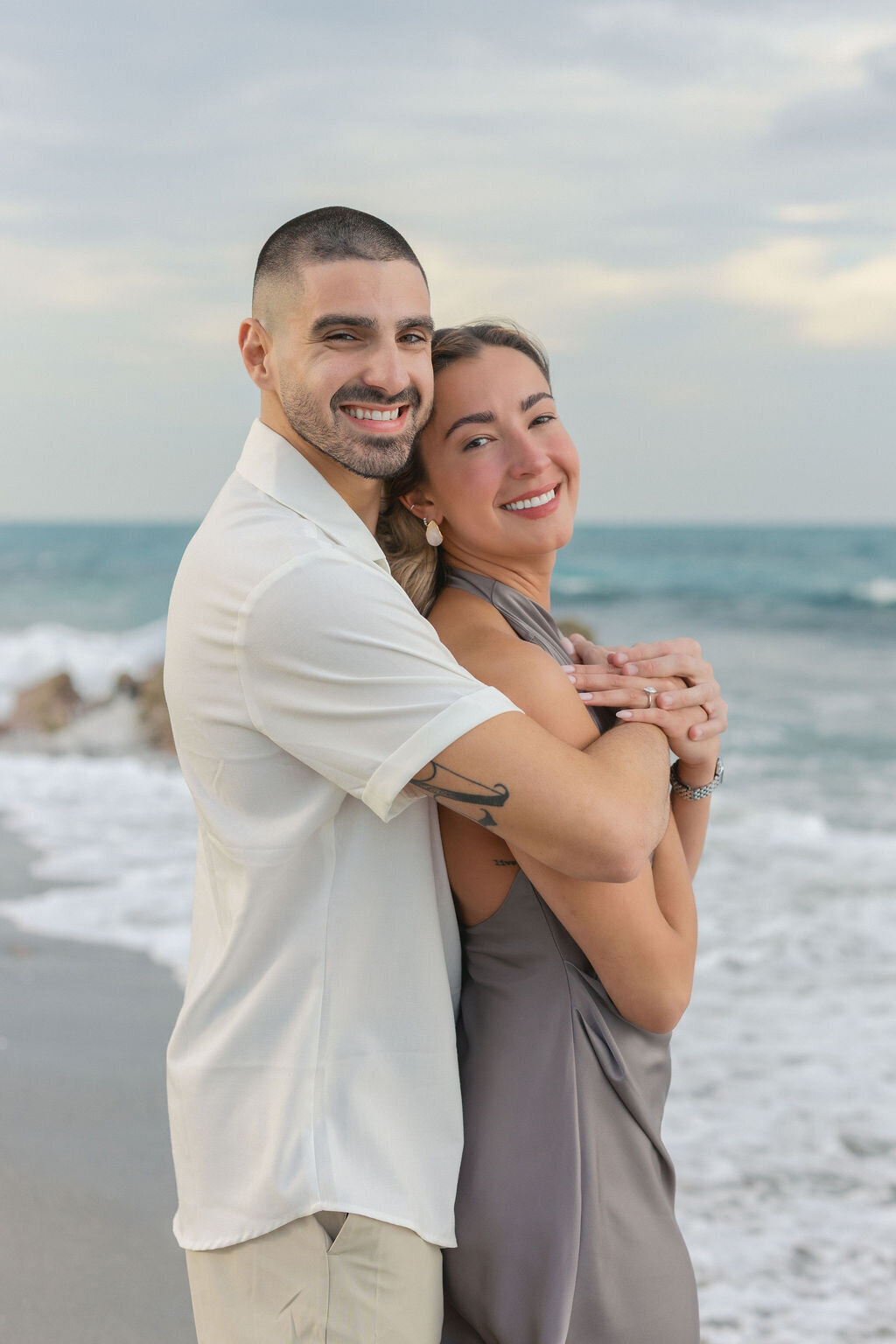 An engaged couple embraces with a bear hug at a south florida beach during sunset.