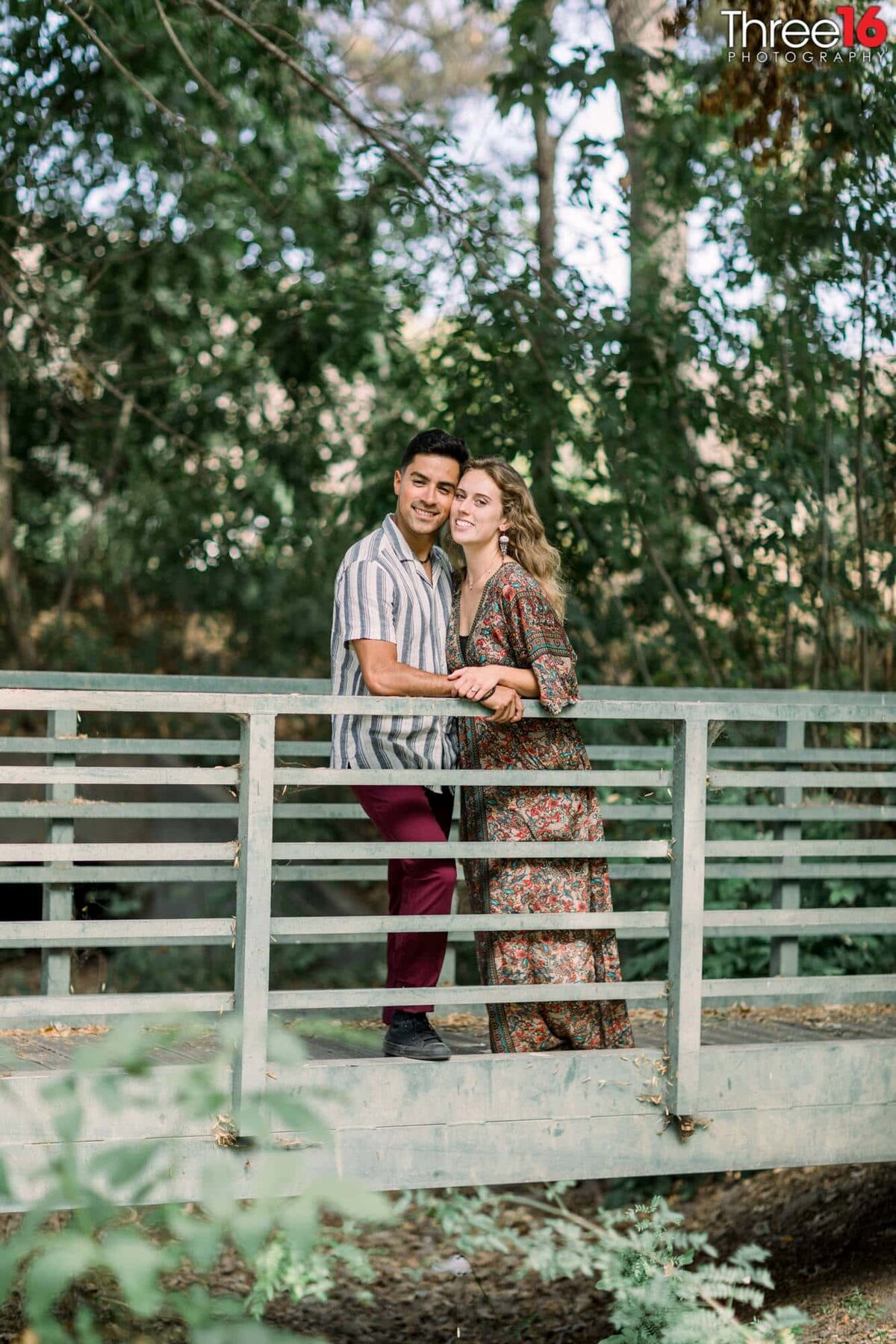 Future Bride and Groom pose for photos leaning against a rail while standing on a bridge that crosses over a creek