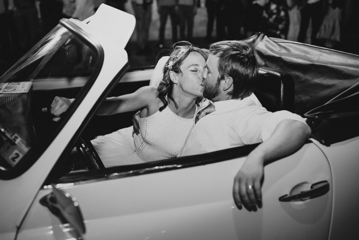 Newlyweds sharing a kiss inside a convertible car, with guests celebrating around them