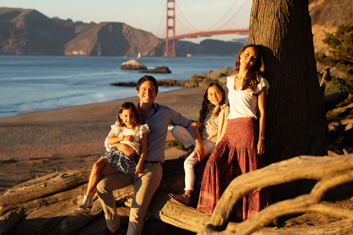 Family with two kids sitting on a tree root at sunset near the beach with the golden gate bridge in the background