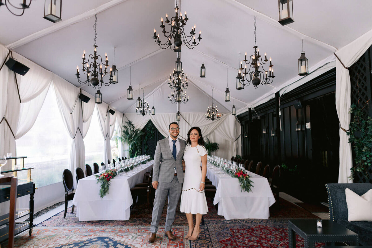 The bride and groom are smiling inside their reception venue, which has candle chandeliers and red flower centerpieces. Image by Jenny Fu Studio
