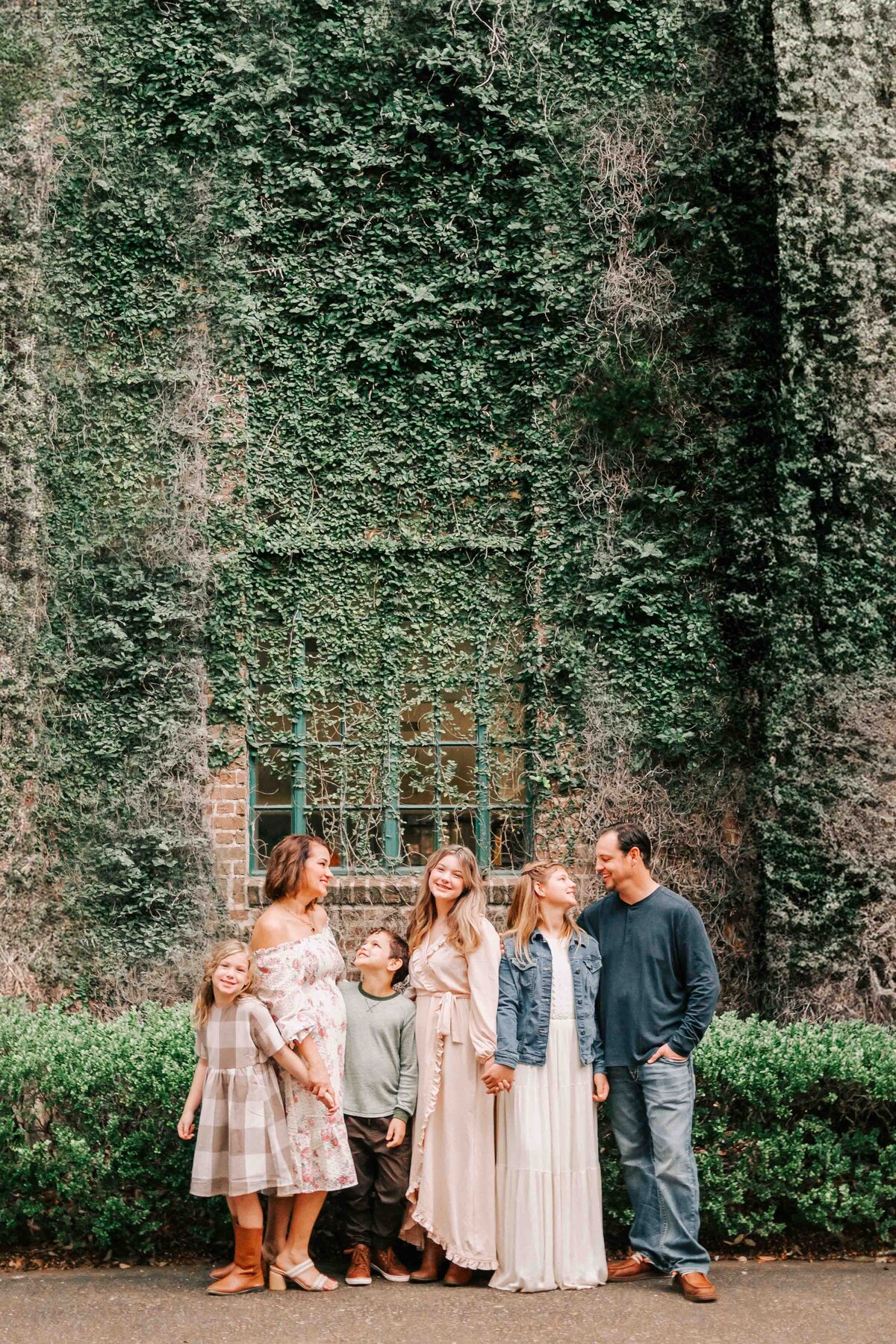 Family stands in front of a green ivy brick wall, all wearing earthy neutral tones.