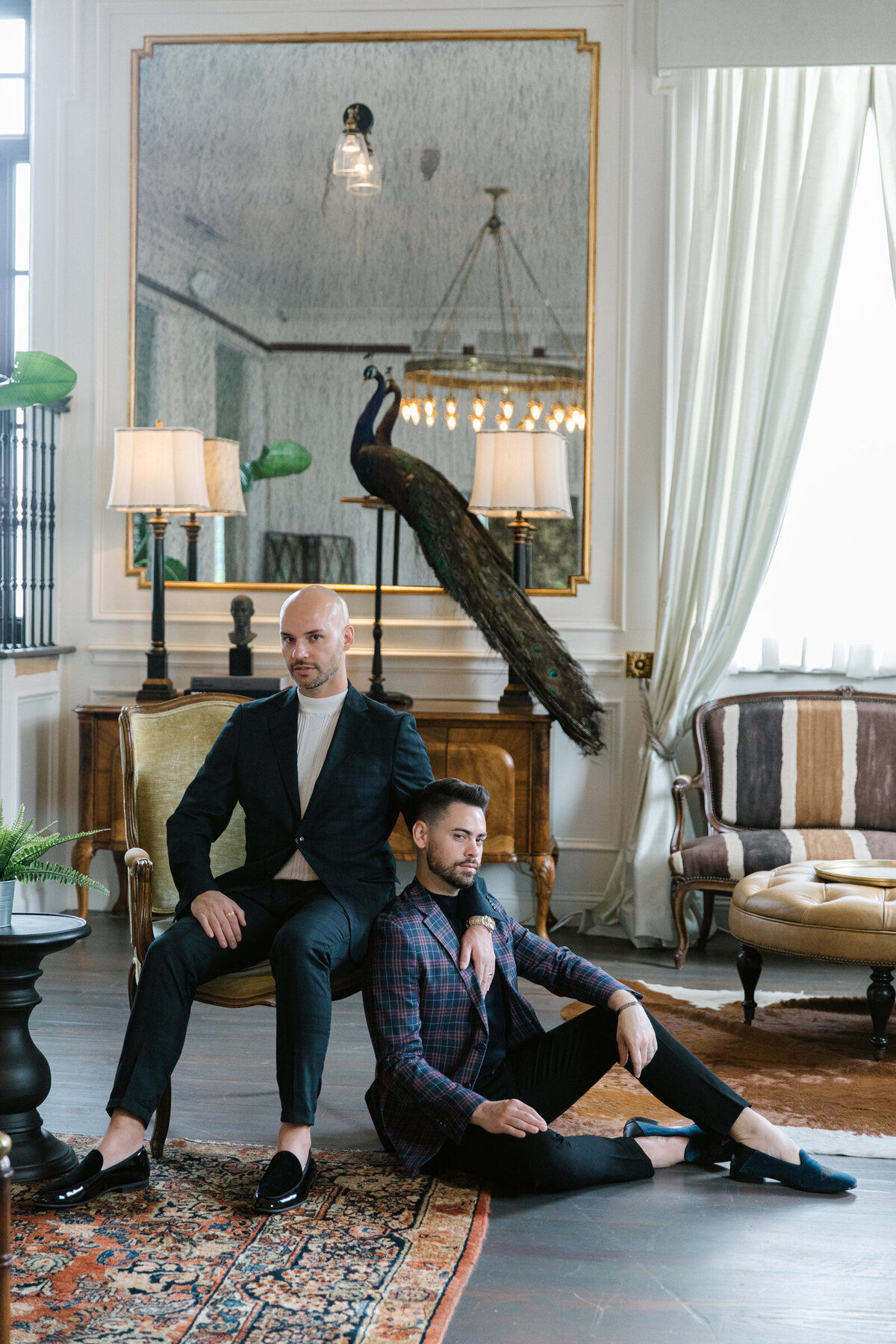 Couple at The Mason in Dallas during their same-sex engagement session, one seated on the floor and the other on a chair, posed in a stylish, royal-inspired portrait.