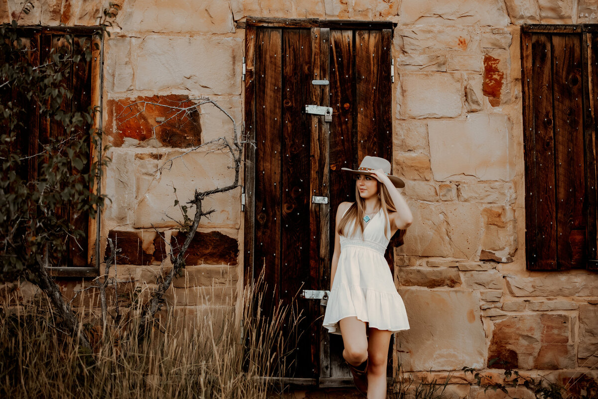 High school senior leaning against a brown wooden door in a white dress and cowboy hat in Boulder, Colorado