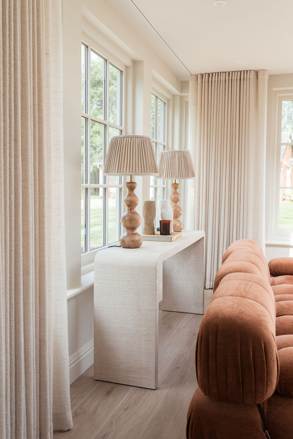 A console table nestled behind a plush, rust orange sofa. Two large neutral lamps sit on the table. Behind the table are lage windows, showing greenery outside.