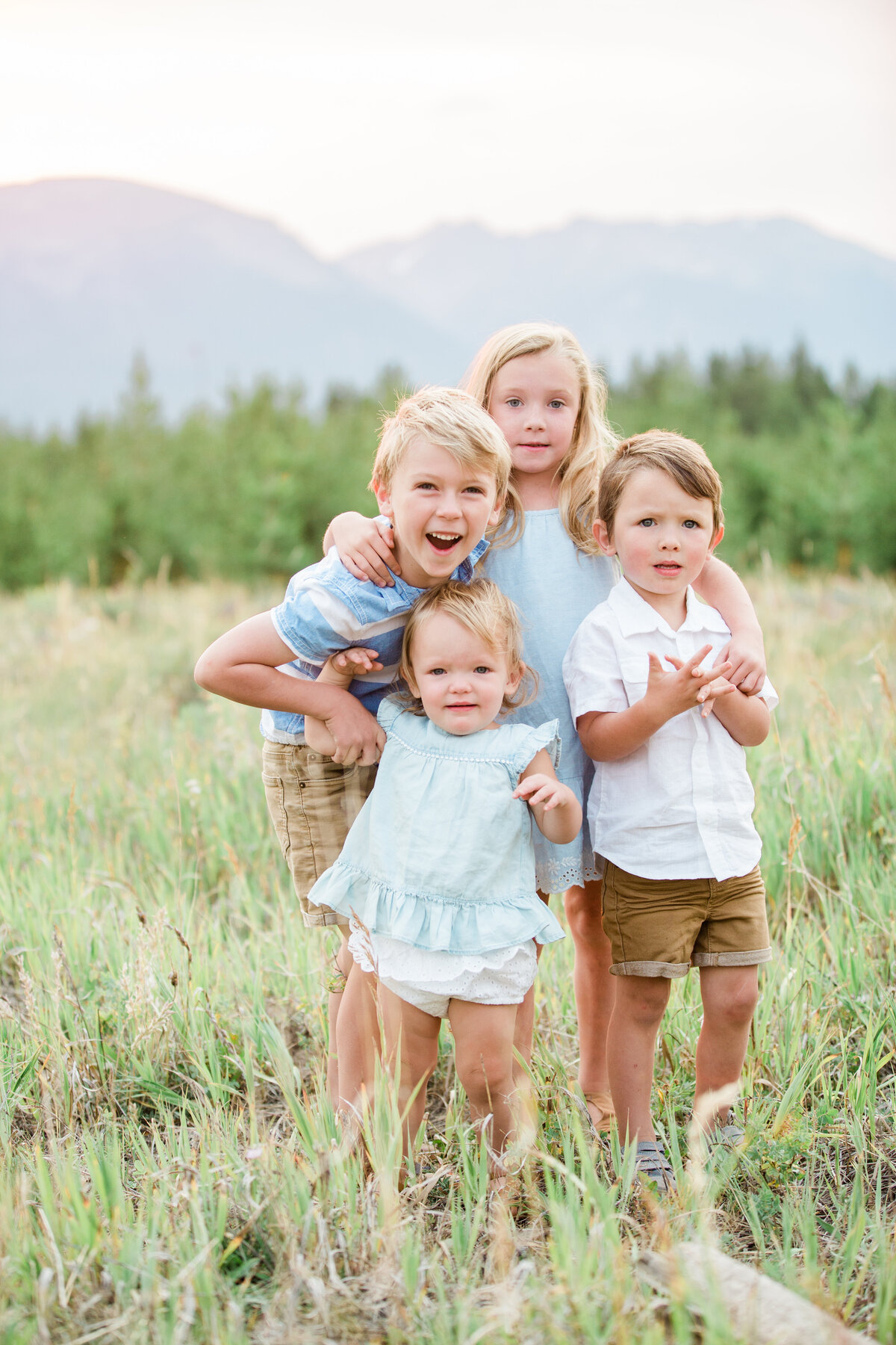Four young siblings are all embraced in a hug. They are in a mountain meadow with high grasses
