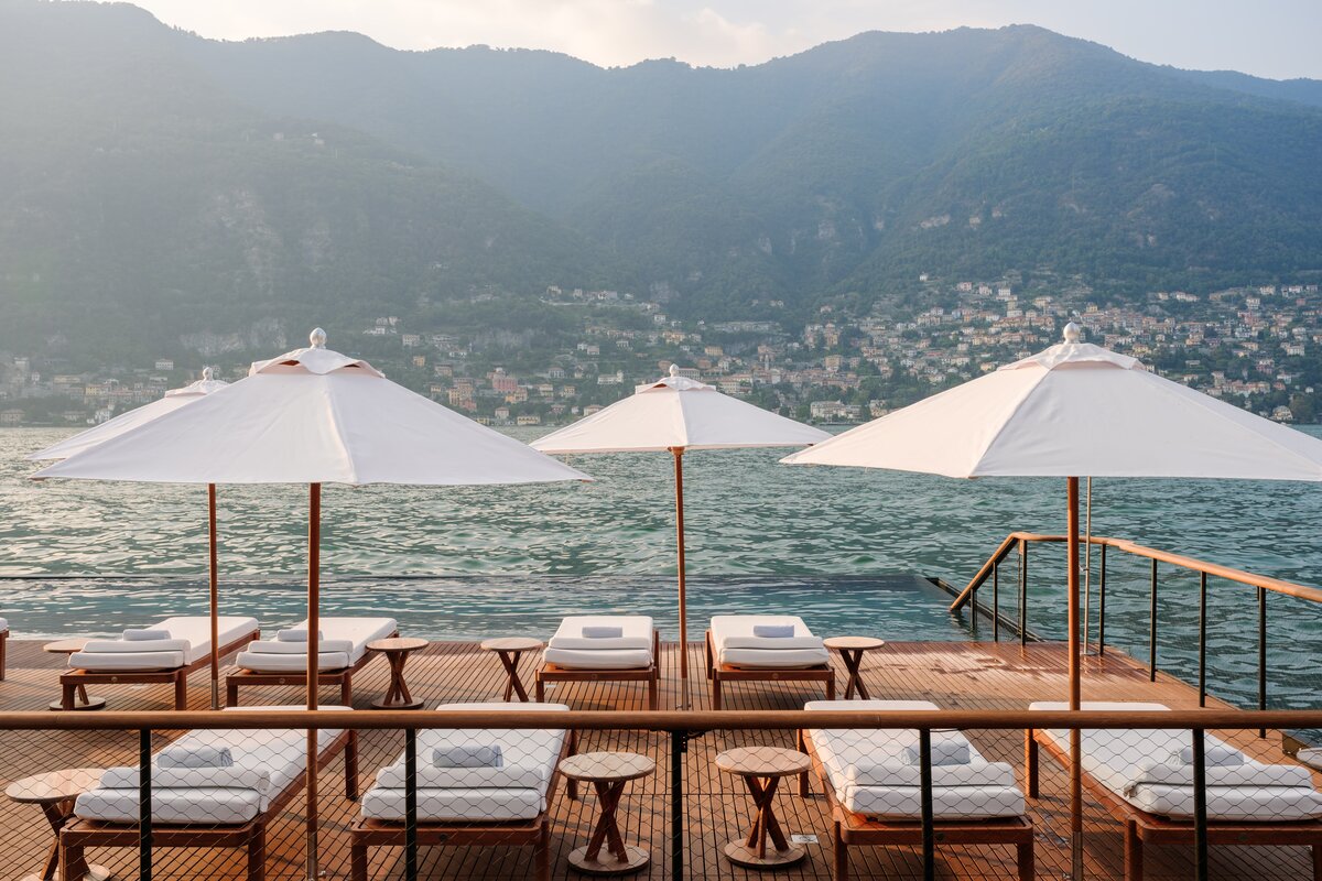 A serene waterfront deck on Lake Como, Italy, featuring plush sun loungers under white umbrellas with breathtaking mountain views. Experience bespoke luxury travel, private villa stays, and high-touch concierge services with Nadia Forte Travel & Events.
