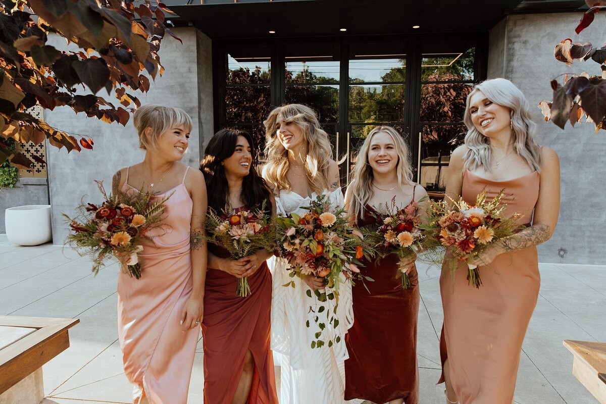 Bridesmaids in blush,  terracotta, peach and brown holding large boho bouquets of burgundy, orange and yellow stand beside the boho bride in a strapless wedding dress with a cascade bouquet of wildflowers and greenery.