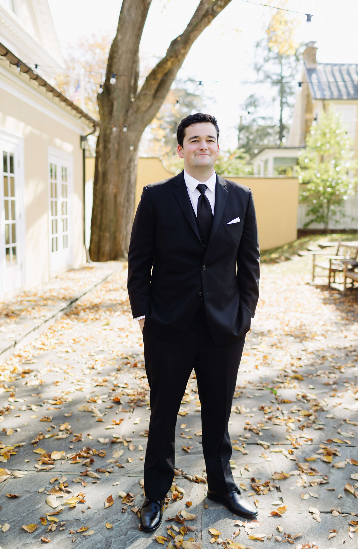 wedding portrait of groom standing on a patio covered in autumn leaves as he stands with his hands in his pockets smiling at middleburg community center