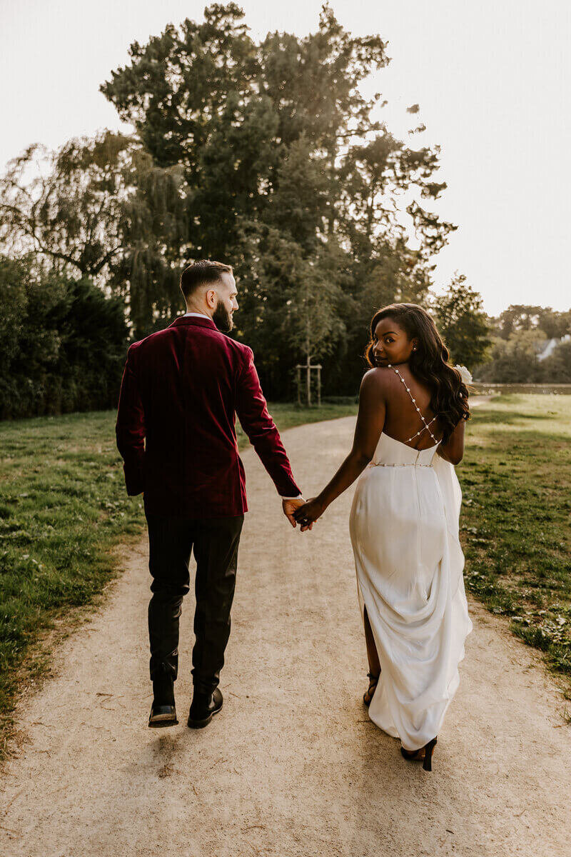 Mariés marchant de dos et se tenant la main en extérieur. La mariée regarde par derrière son épaule. Photo prise par Laura, photographe mariage en Vendée.