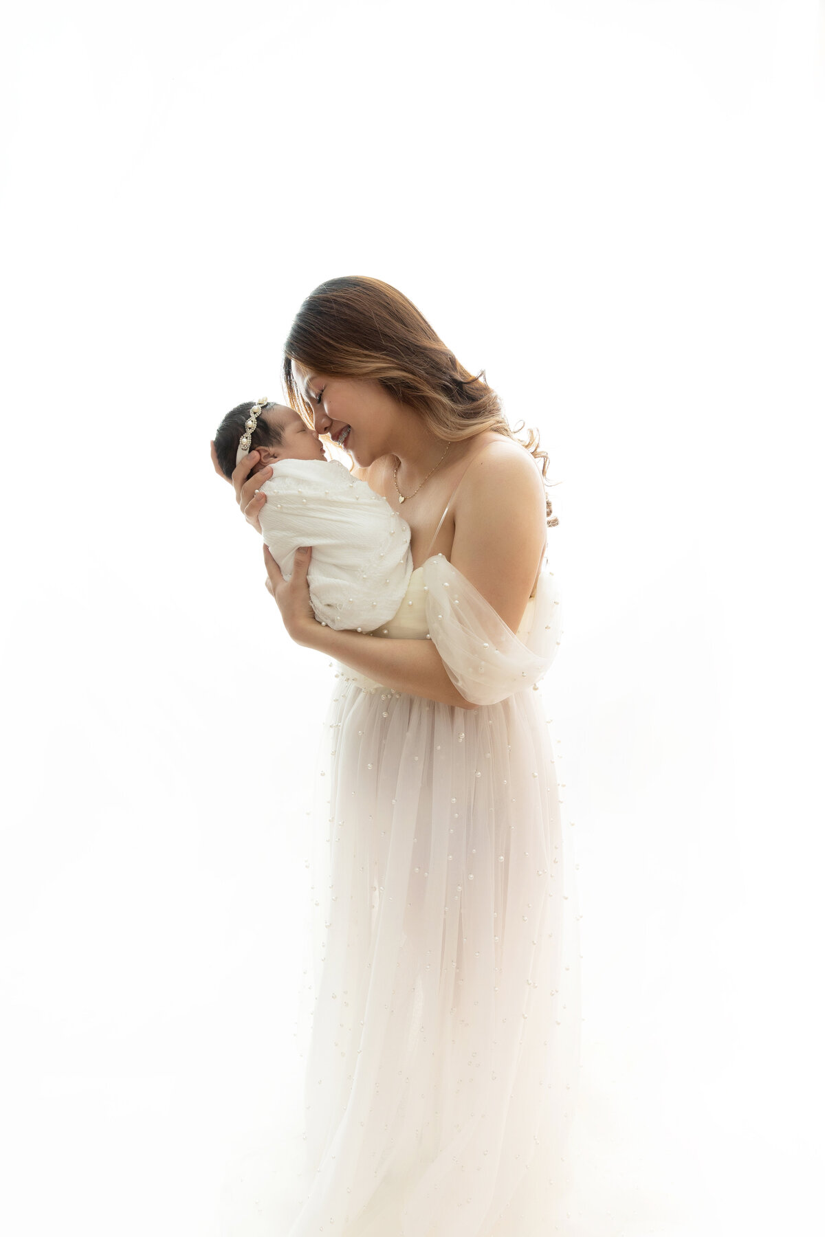 A happy mother nuzzles noses with her sleeping newborn baby daughter while standing in a studio in a white dress