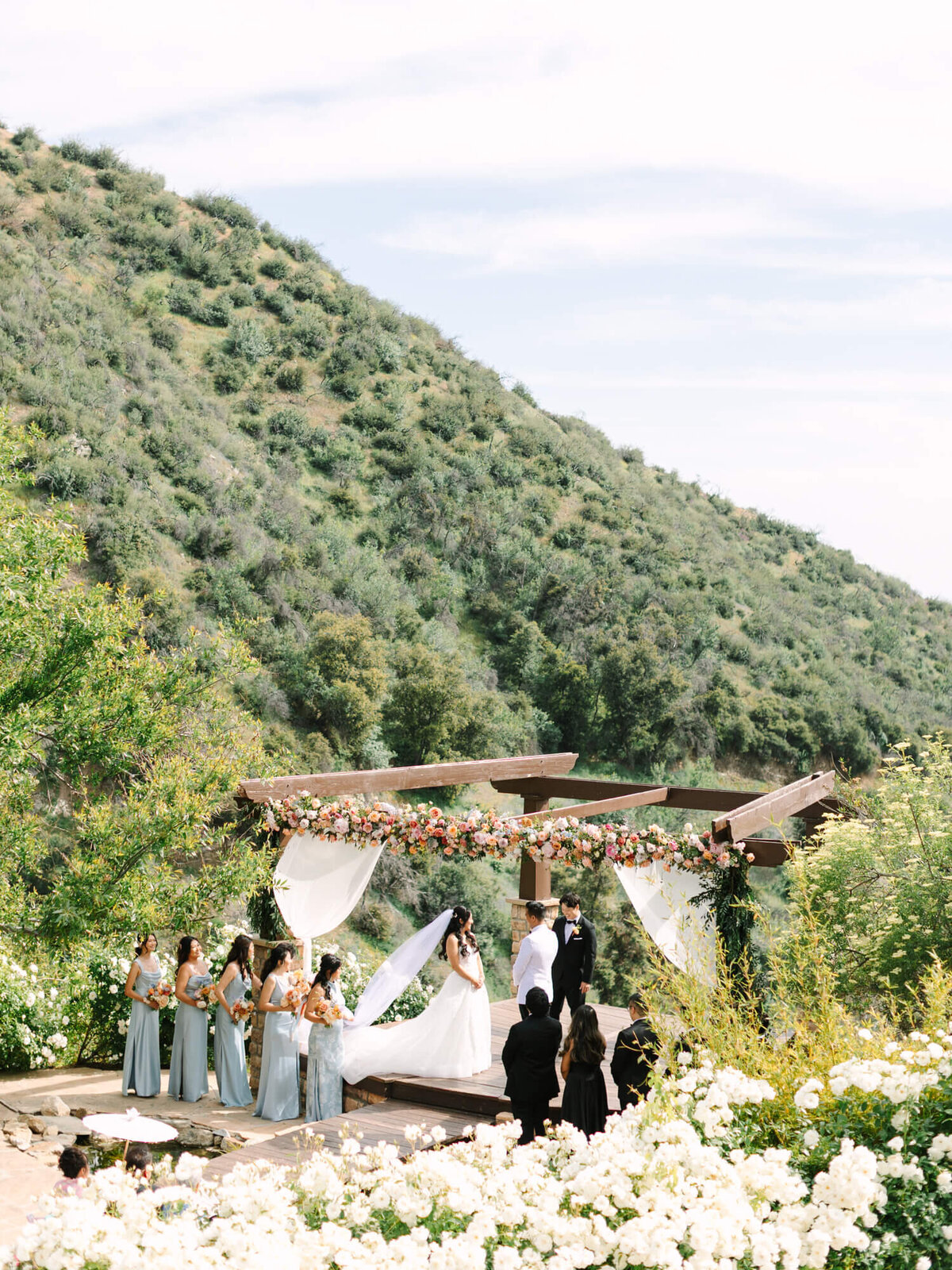 Elegant outdoor wedding ceremony set against a lush hillside at Serendipity Garden Weddings Venue. A bridal party in light blue stands beside the couple under a flower-adorned wooden pergola.