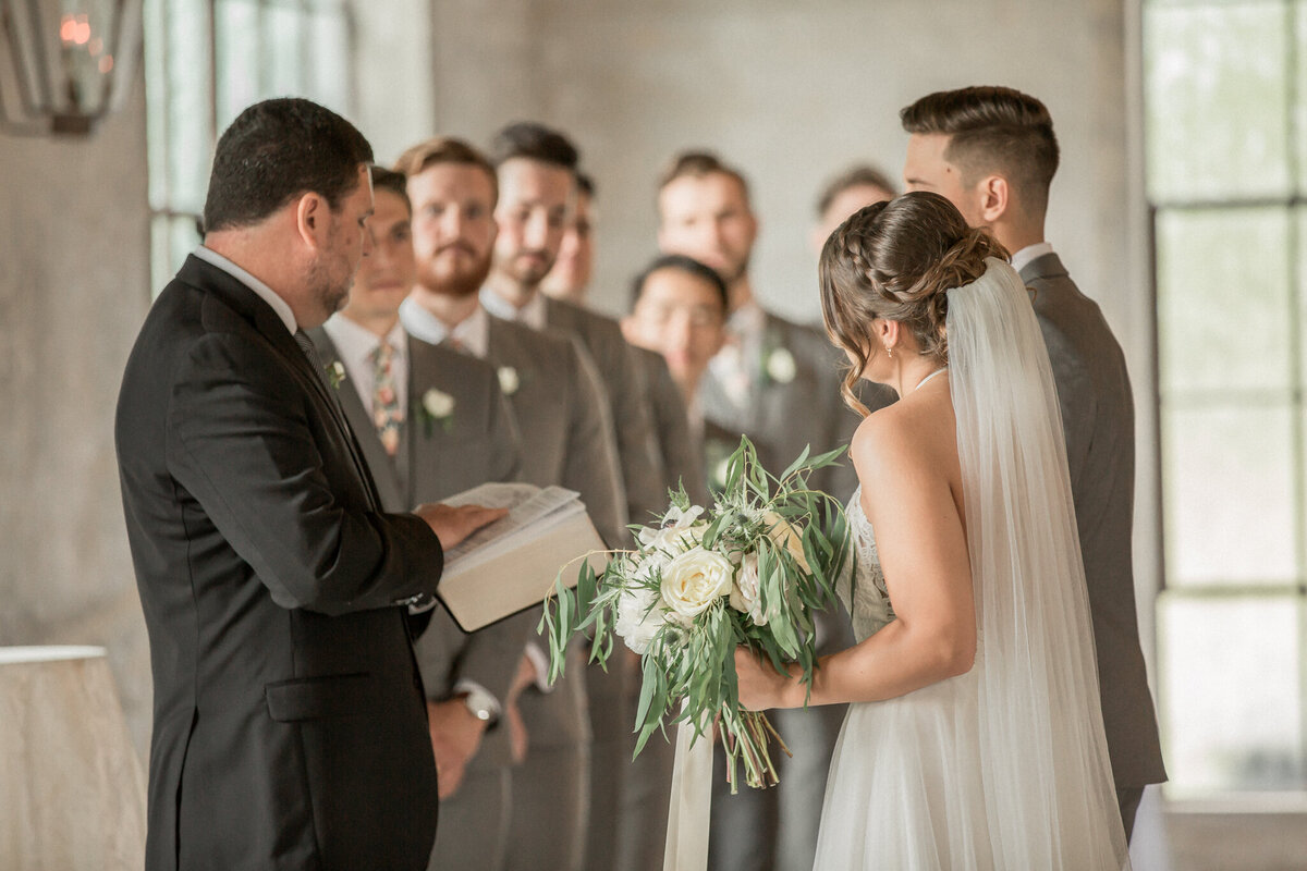 The bride and groom stand close to their pastor while the groomsmen look on