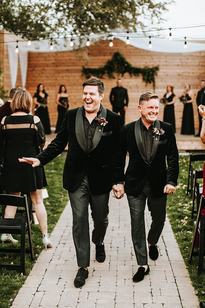 Two grooms wearing black tuxedos walk down the aisle smiling holding hands at their wedding while waving at guests.