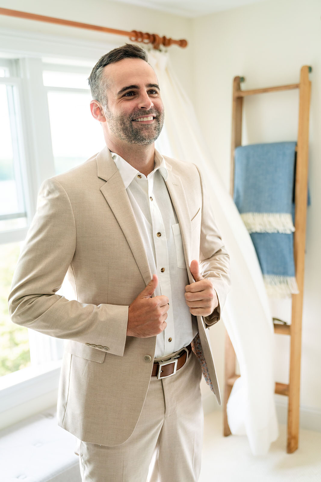 A cheerful man buttoning up his beige suit jacket, preparing for a special occasion in a sunlit room with a ladder blanket rack in the background.