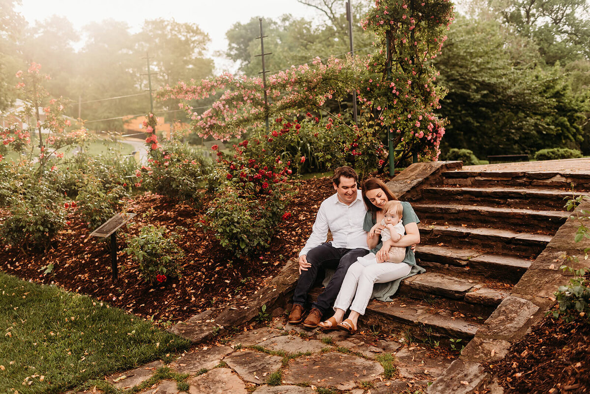 mom, dad, and one year old son sitting on steps in a rose garden.  Son is sitting on mom's lap, while they all laugh and play.  Behind them is a hill of rose bushes, and golden morning light.  Photo taken by Philadelphia family photographer, Kristi