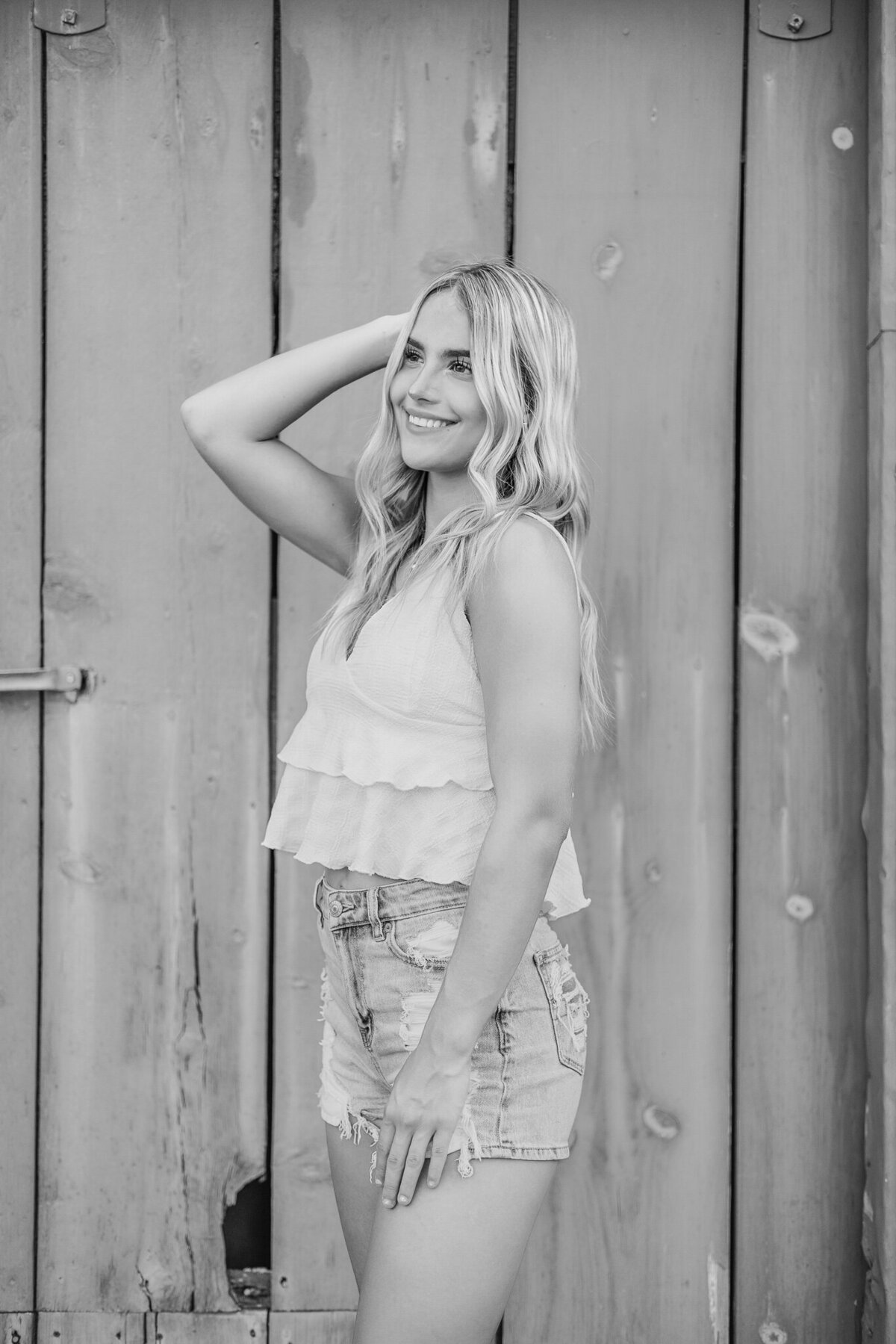 black and white photo of a high school senior female, standing in front of a door, standing to the side, looking off camera smiling, with hand at her side and the other in her hair, wearing blue jean shorts, and spaghetti strap shirt.