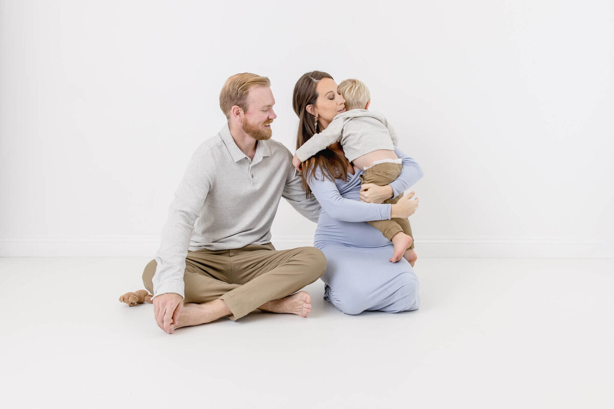 A mother in a light blue dress holds and kisses a young child in a gray shirt and pants. A father, wearing a light gray shirt and beige pants, sits beside them, smiling. The family is seated against a plain white background.