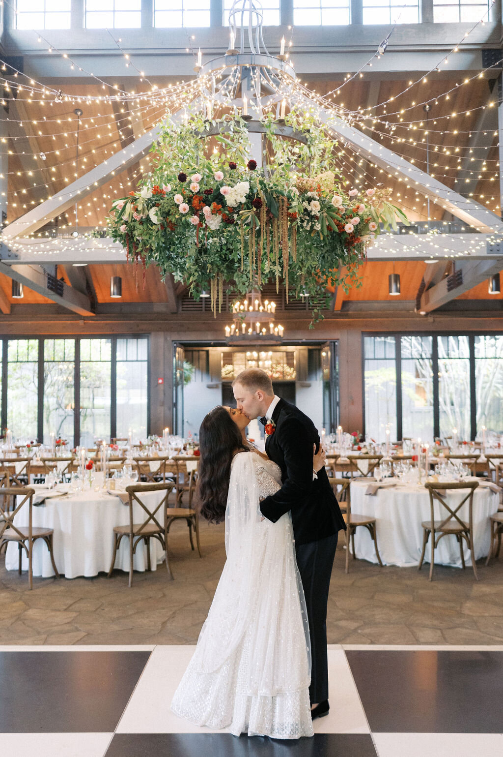 Bride and Groom share a kiss in front of wedding reception with large hanging chandelier floral arrangement. 