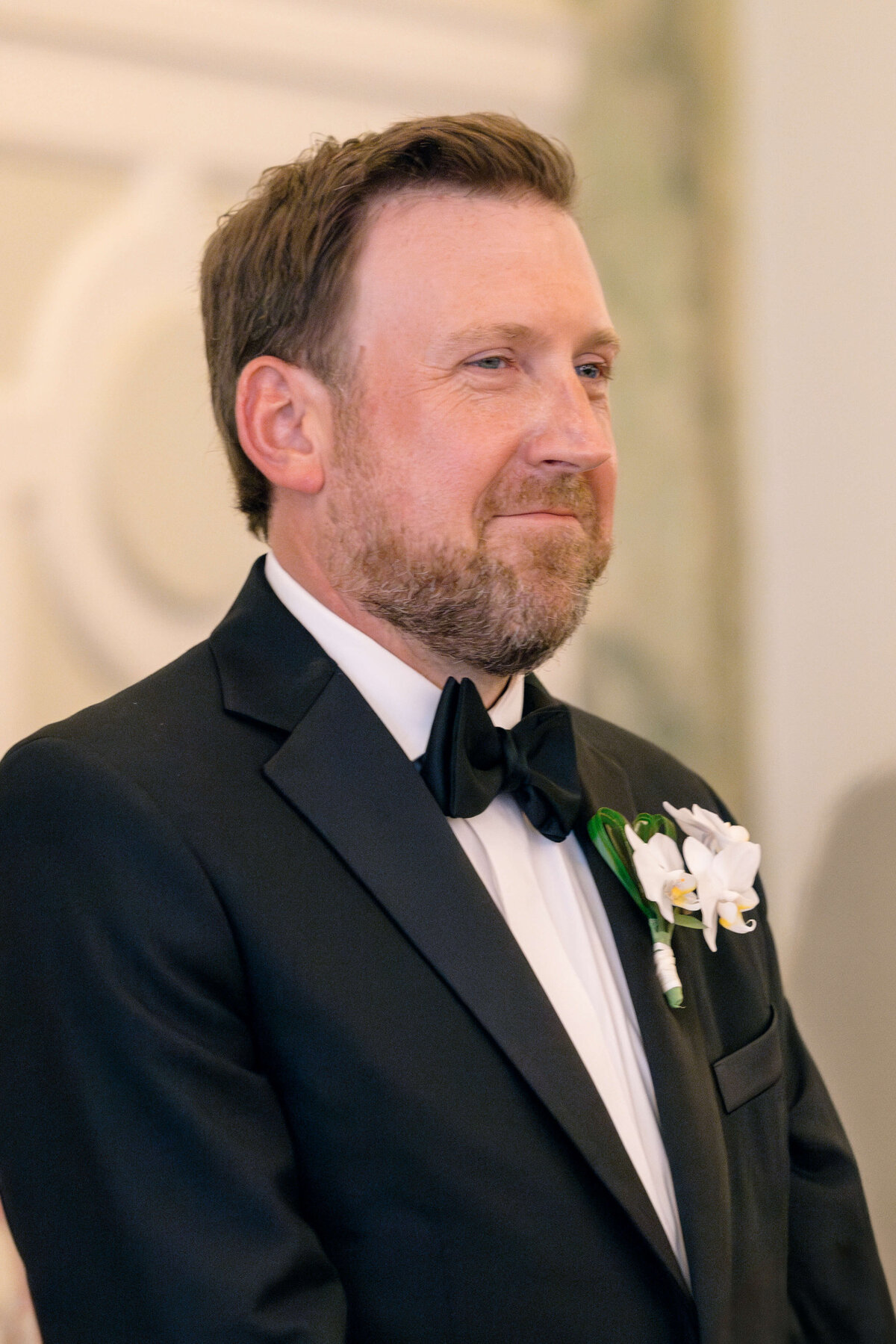 A man with short hair and a beard is wearing a black tuxedo and bow tie, standing with a smile. A white flower boutonniere is pinned on his jacket. The background is softly blurred with a light-colored wall.