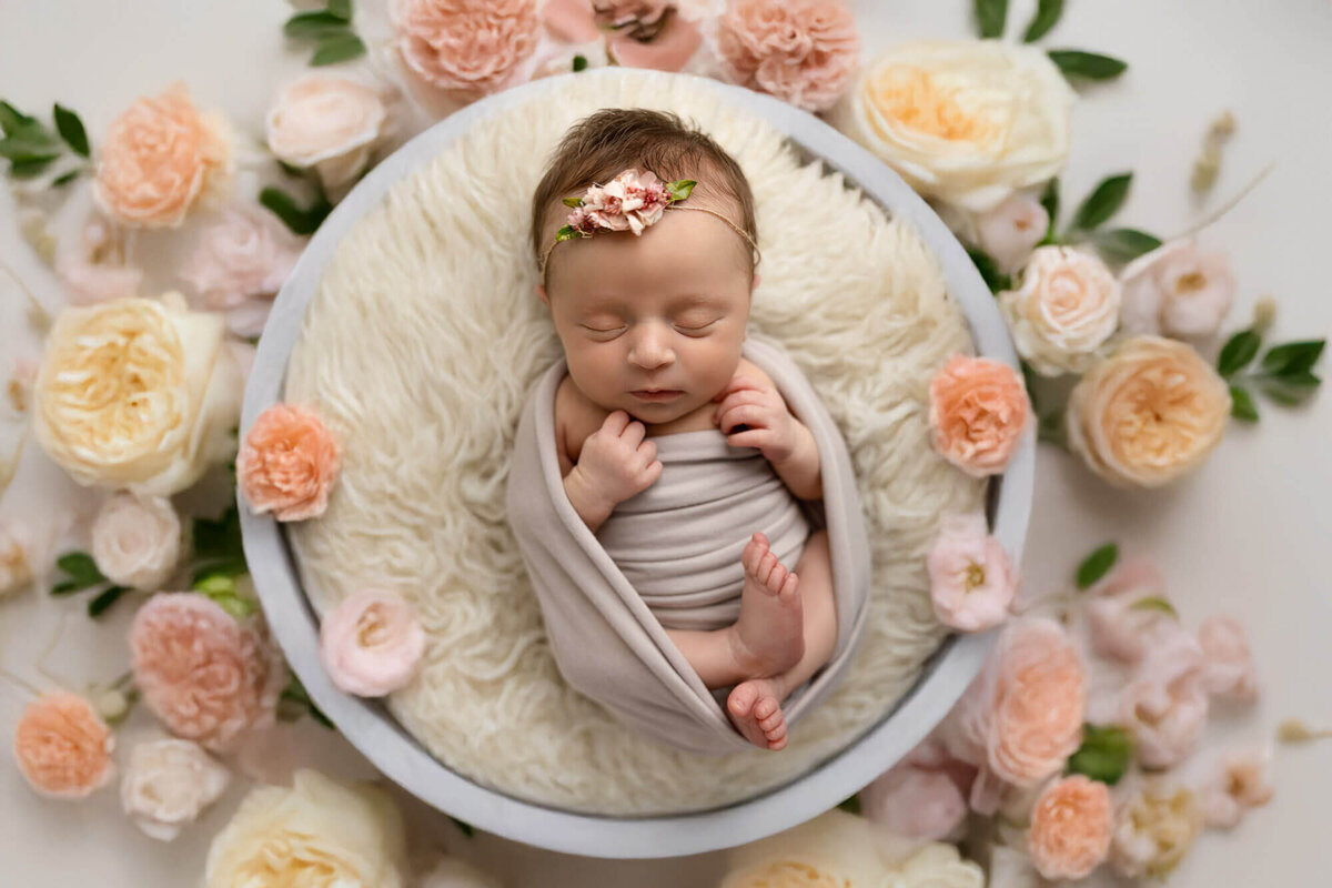 Newborn baby girl wrapped in cream swaddle surrounded by flowers during studio session