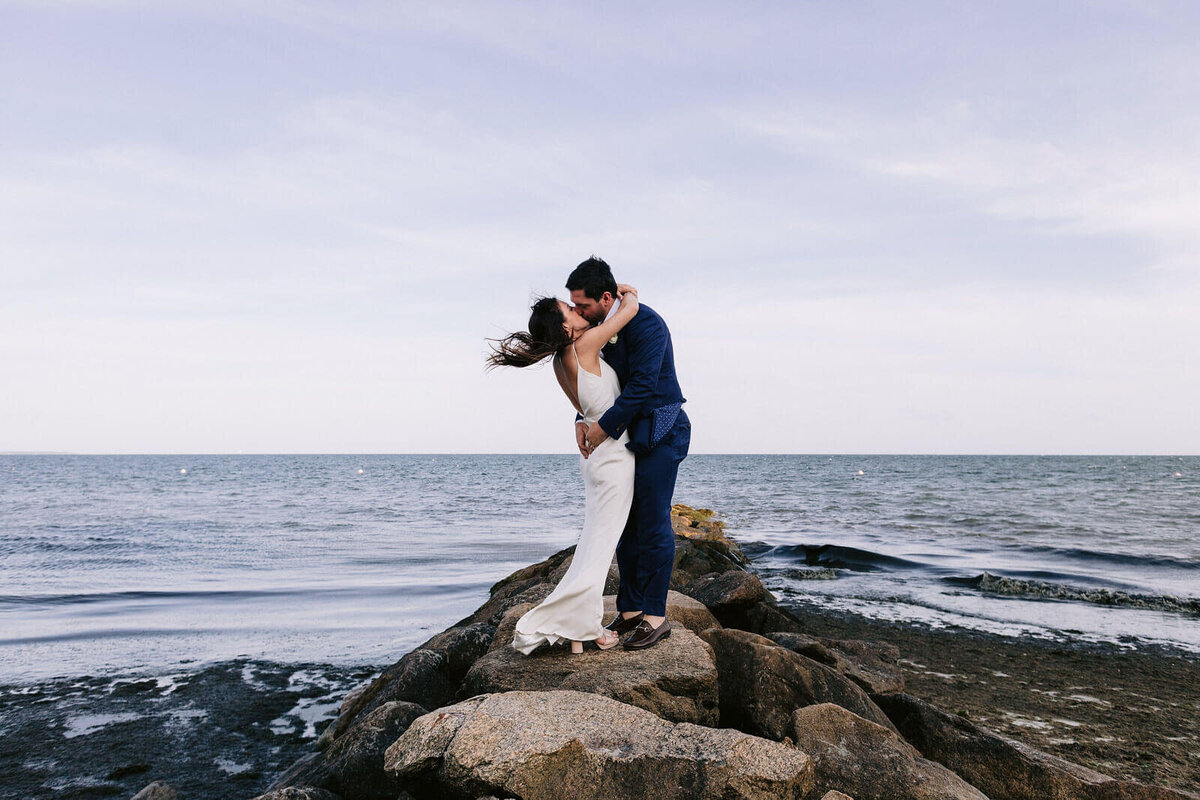 The bride and the groom are kissing each other atop a rock on a seashore in Cape Cod, MA, with the ocean in the background.