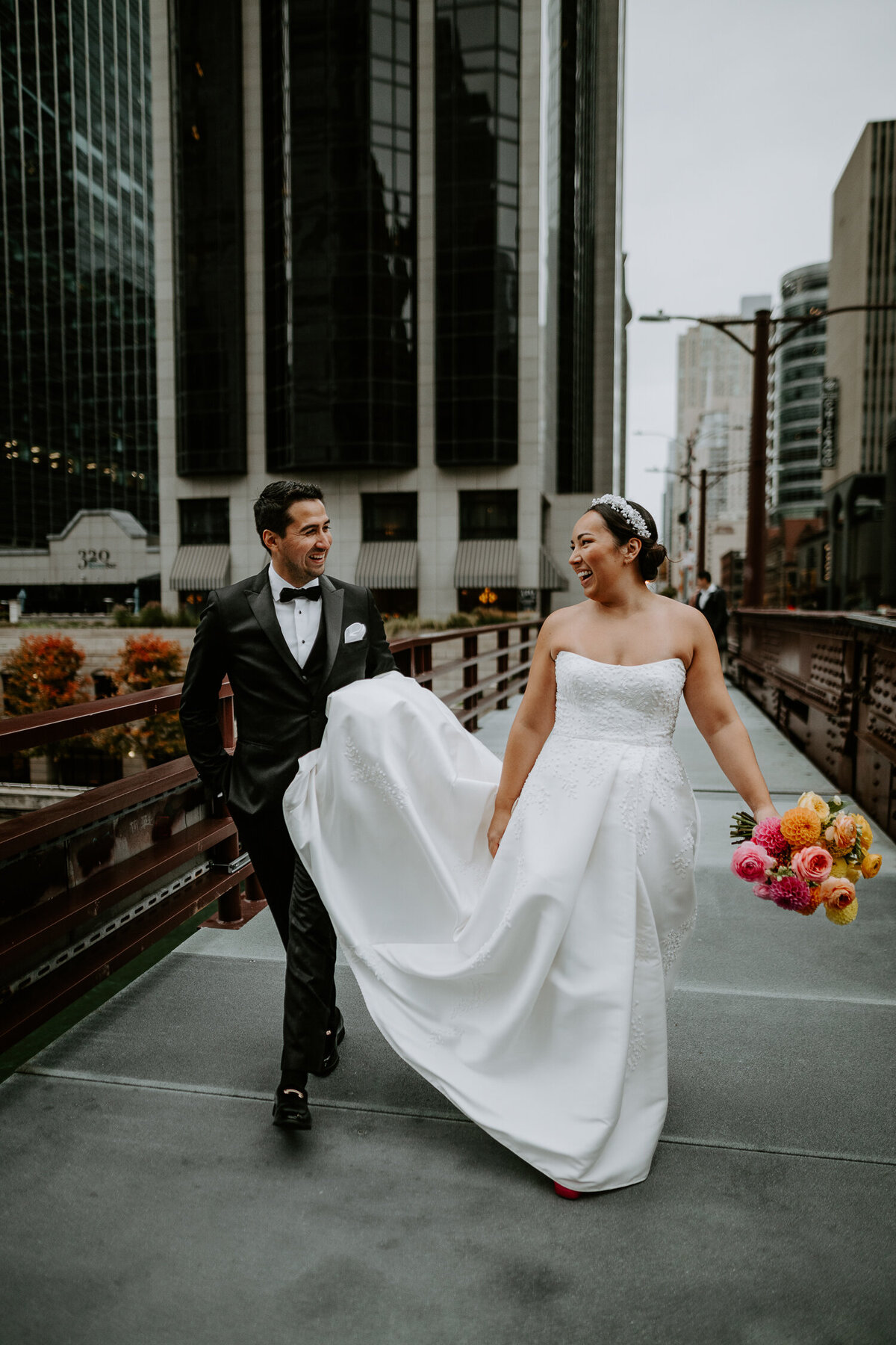 A groom carries his Bride's dress as they walk over the bride of the River Walk in Chicago.