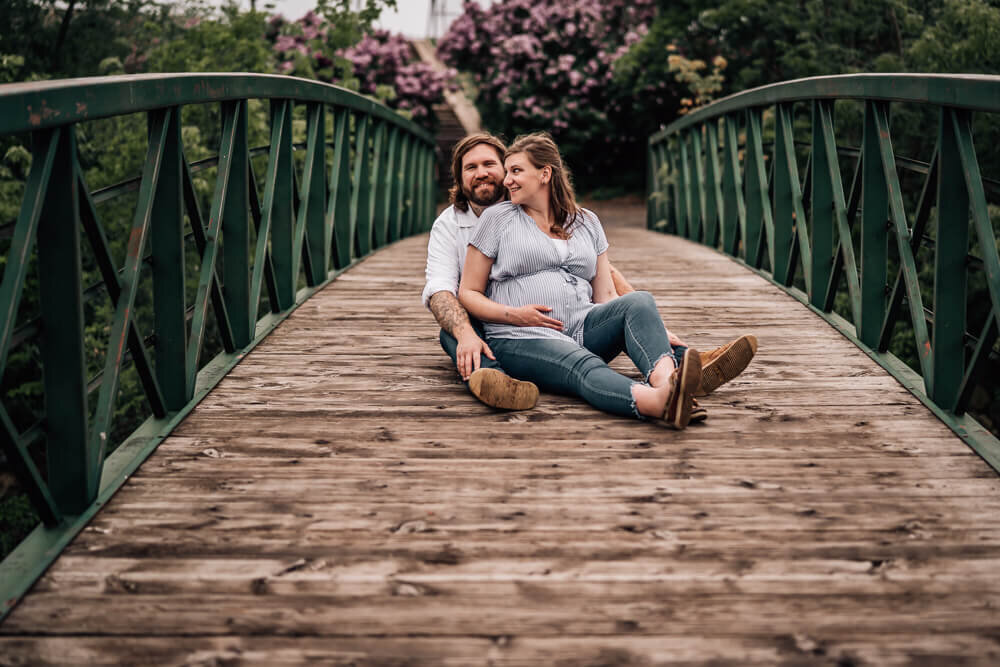 A couple expecting their first child soon, sit on a bridge with blooming lilacs in the background at Boom Island Park in Minneapolis, MN.