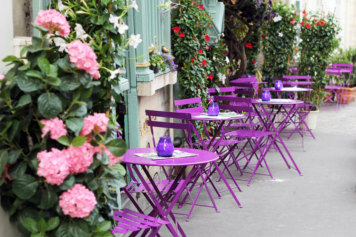 Sidewalk café with vibrant purple tables and chairs, adorned with purple candle holders. Lush pink and red flowers along the green storefront add a cheerful ambiance to the outdoor seating area.