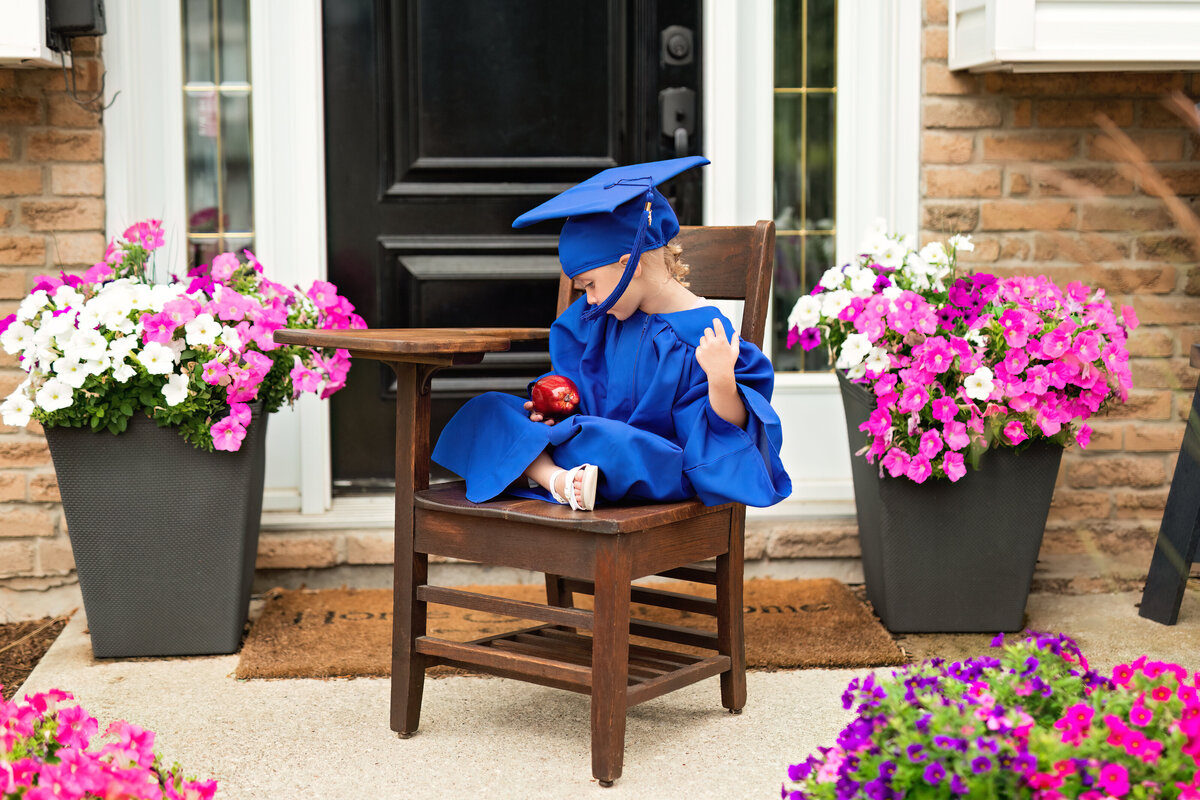 Quiet milestone moment of a little girl in graduation attire studying an apple—symbolizing knowledge, growth, and grace.
