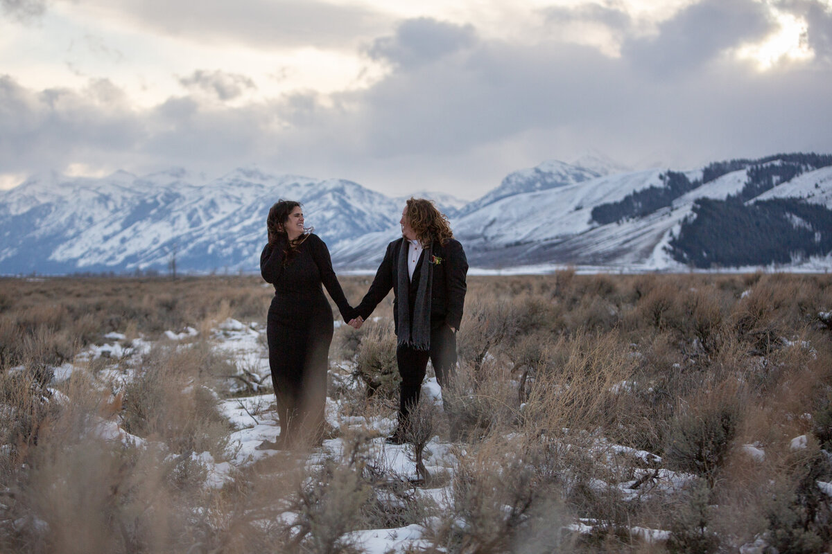 Two brides wearing black walk through a snowy meadow at sunset during their elopement in Grand Teton National Park.