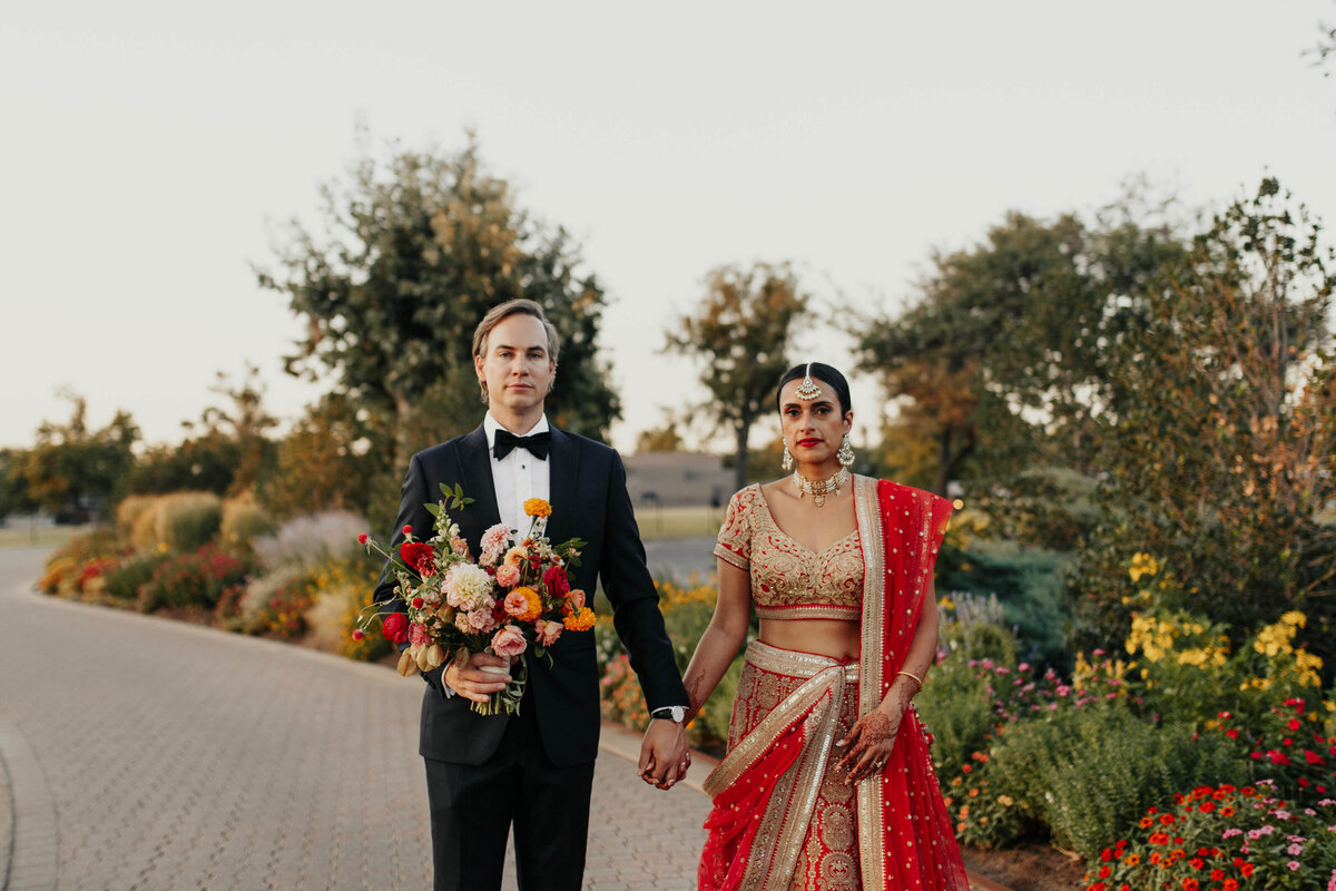 A couple stands, holding hands. The woman is in a traditional red Indian dress and the man is in a tuxedo, holding a bouquet of red and yellow flowers.