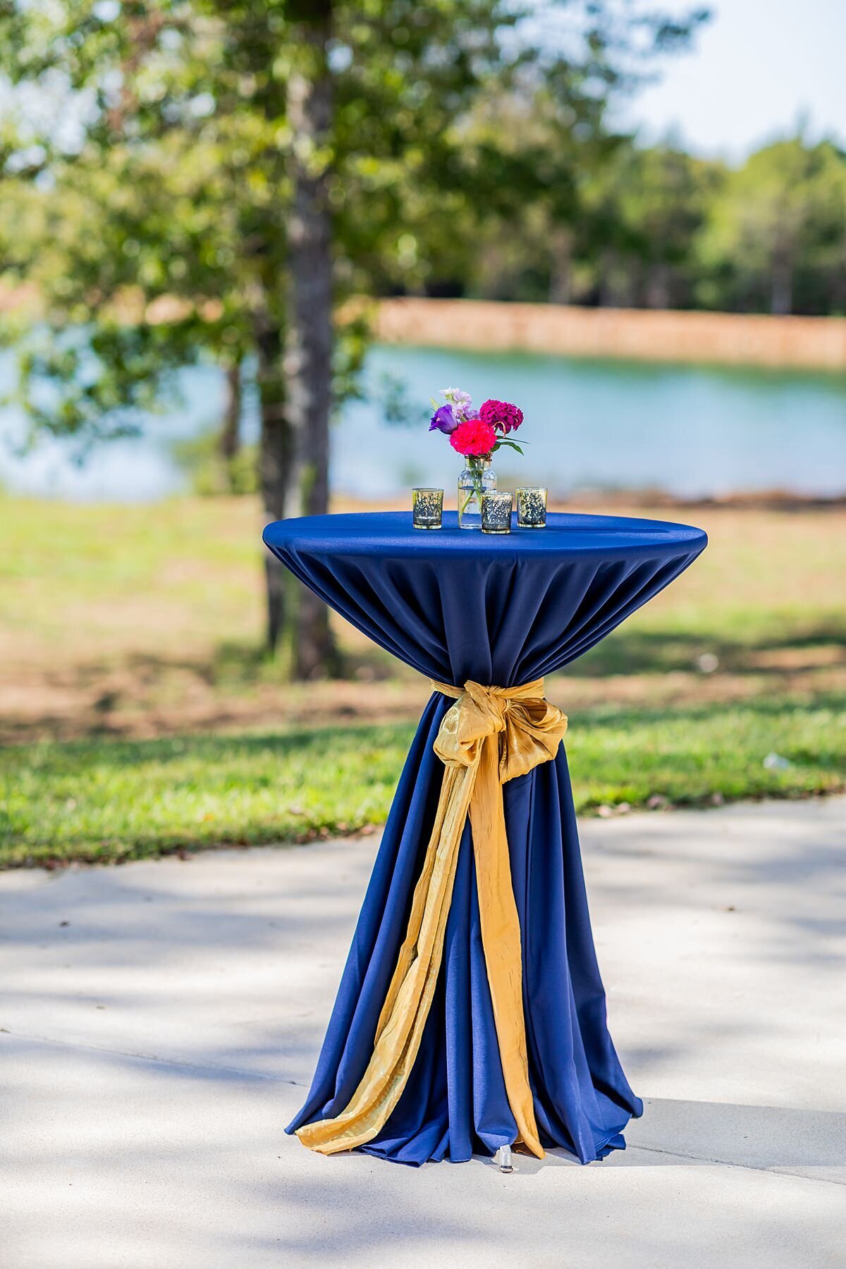 Cocktail table with a royal blue table cloth tied with a gold sash and topped with a bud vase of pink and purple flowers surrounded by gold votives sits lakeside at Sugarfoot