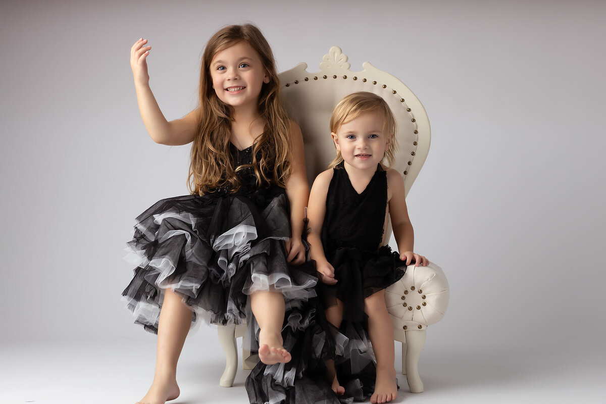 Two girls in black and white dresses, one waving and the other smiling, sitting together in a vintage chair.