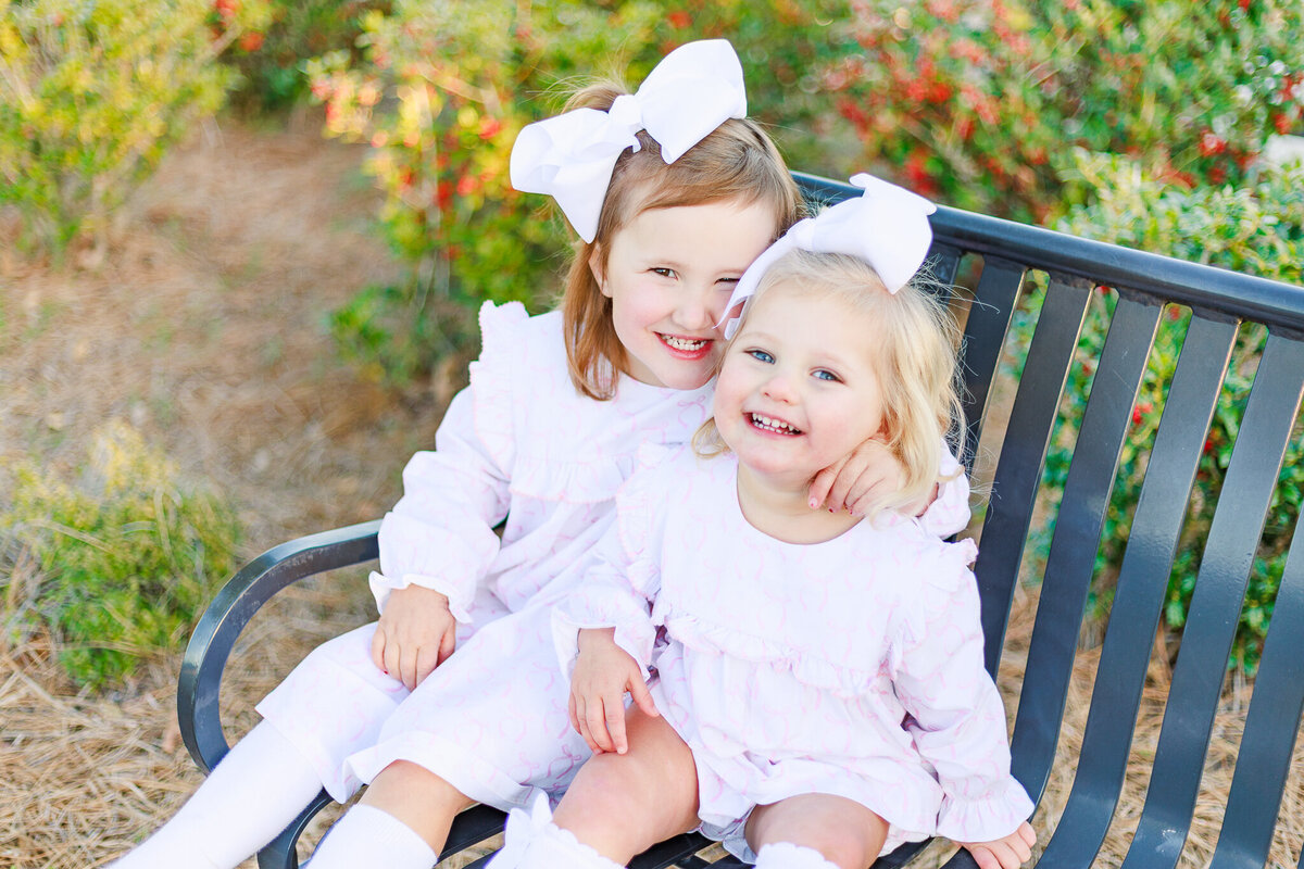 Two young sisters with big white bows and matching pastel dresses sit close together on a park bench, laughing joyfully. Their sweet embrace and radiant smiles capture the warmth of childhood and sisterly love in Birmingham, Alabama.