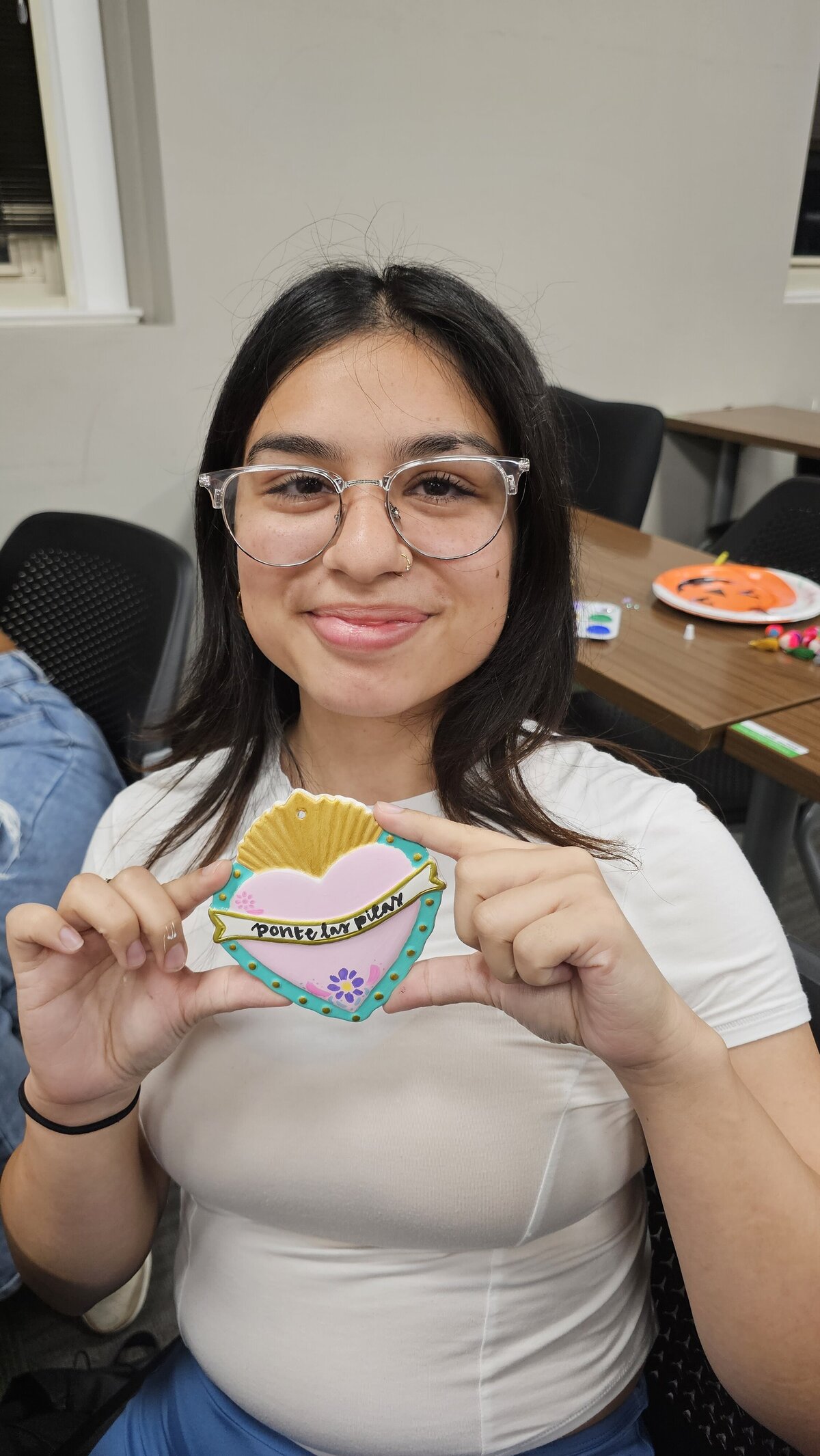 The artistic prowess of a young girl seated on a white plastic chair as she proudly presents her final piece from the workshop at Soleil Vida Studio. Dressed in a white t-shirt with reading glasses as a stylish accessory, she crafted a heart in vibrant blue, red, and yellow, adorned with a poignant inspirational phrase