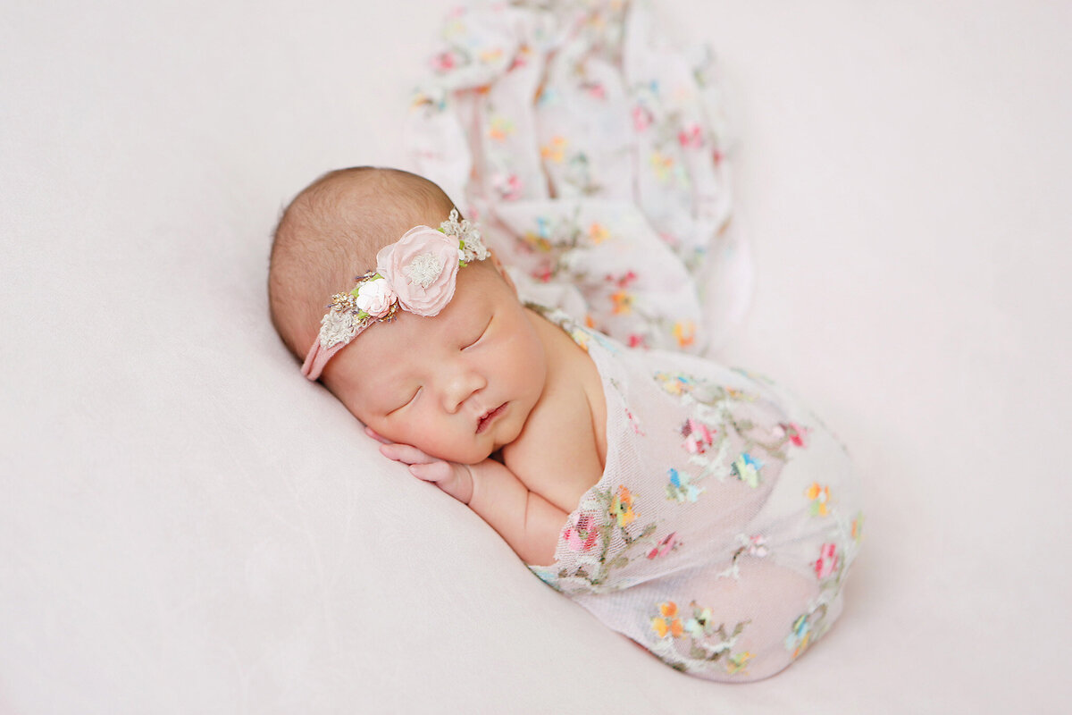 Baby girl laying on a soft pink backdrop asleep wearing a pink flower headband and pink flower swaddle photographed by Los Angeles newborn photographer Elsie Rose Photography