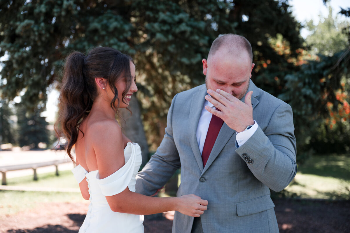 bride and groom standing in front of tree and groom wiping his eyes after emotional first look