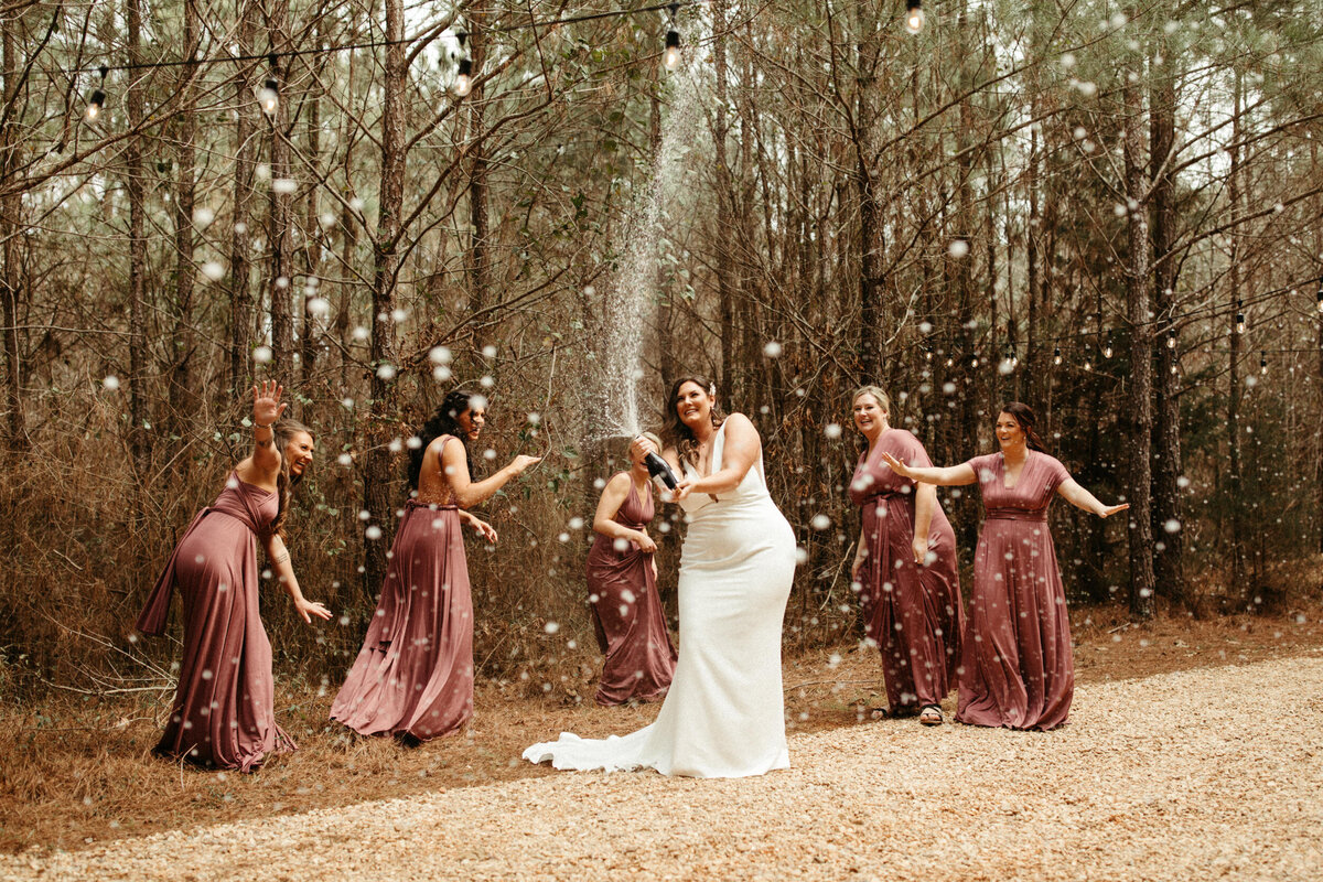 Bride popping champagne as her bridesmaids laugh and run away in the woods