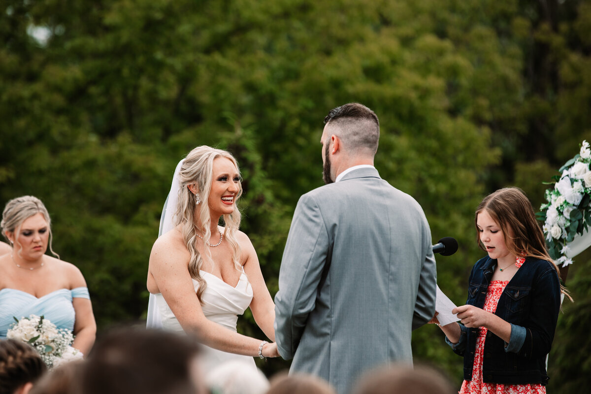 A bride and groom hold hands while smiling at each other during a reading at their ceremony