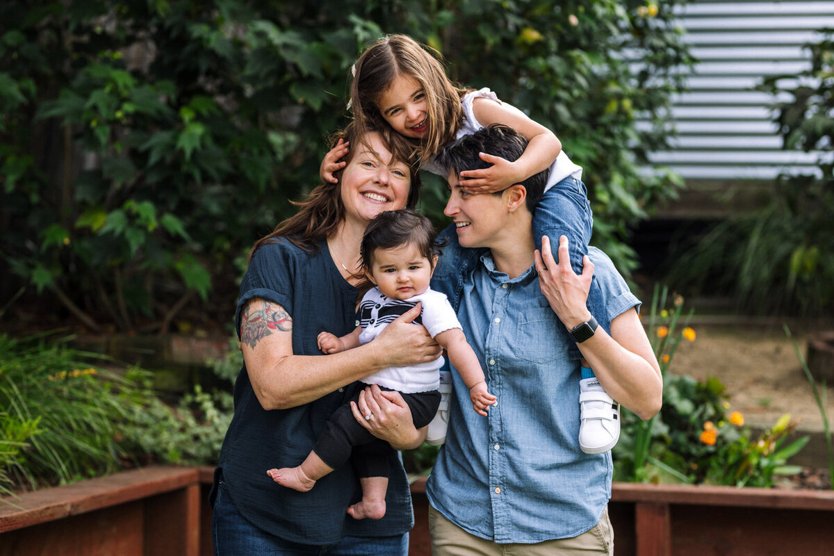 two moms holding toddler and baby daughters smiling and interacting with each other in their Oakland backyard