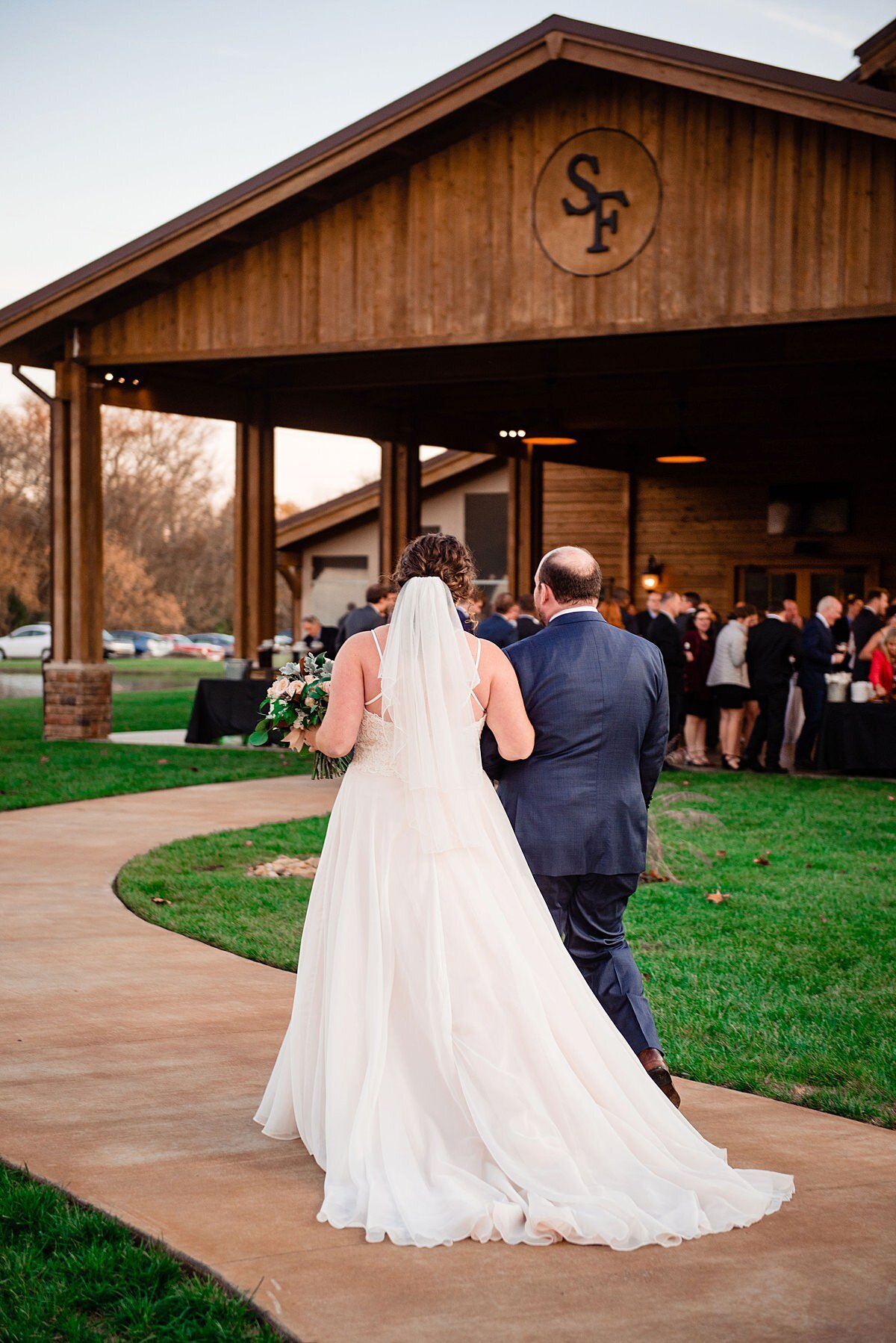 The bride, wearing a long lace wedding gown and veil walks hand in hand with her groom dressed in a dark navy suit towards their guests celebrating their fall wedding at Sycamore Farm