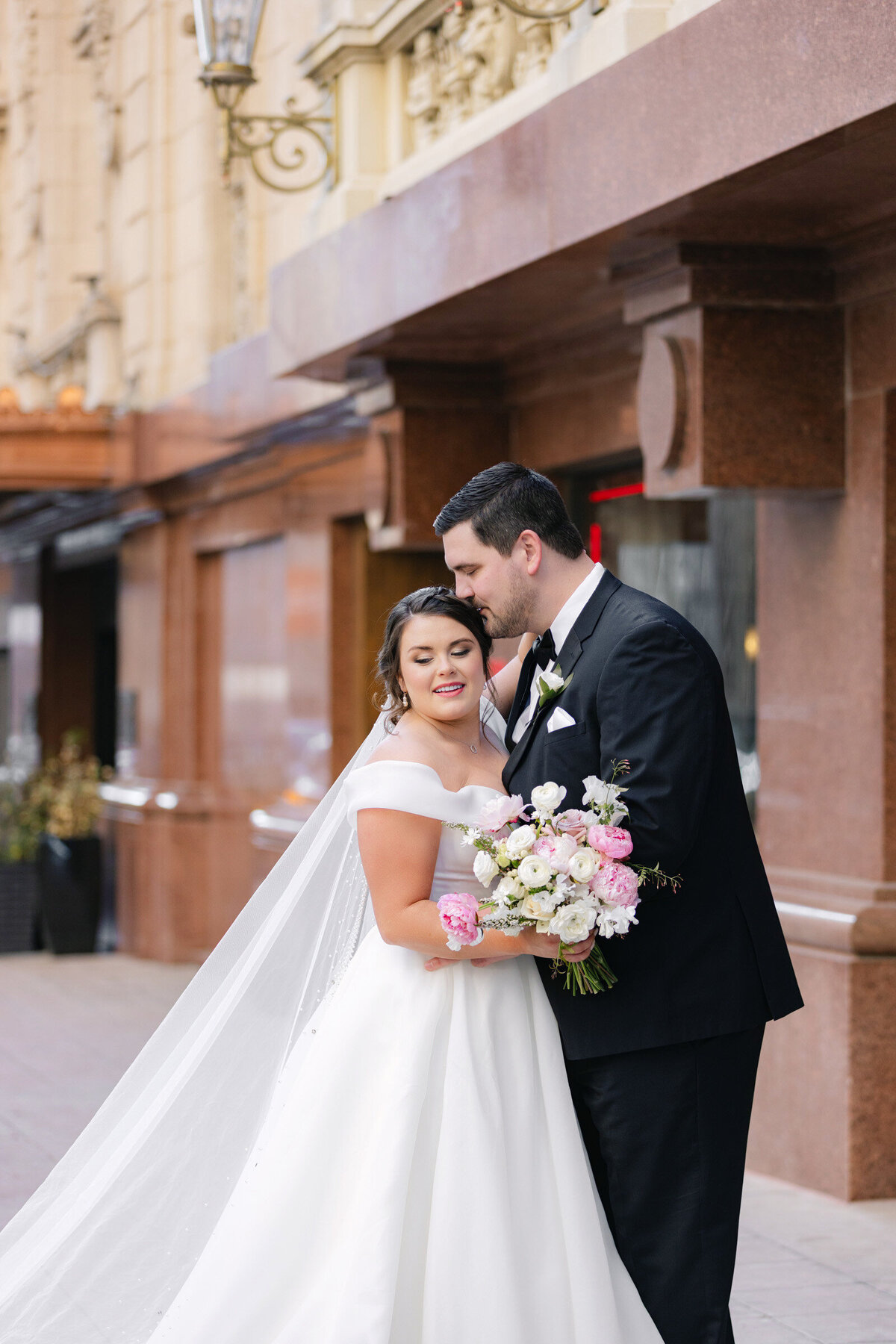 bride and groom outside The Adolphus in Dallas, with the groom kissing the bride on her head, capturing a tender and romantic wedding moment.