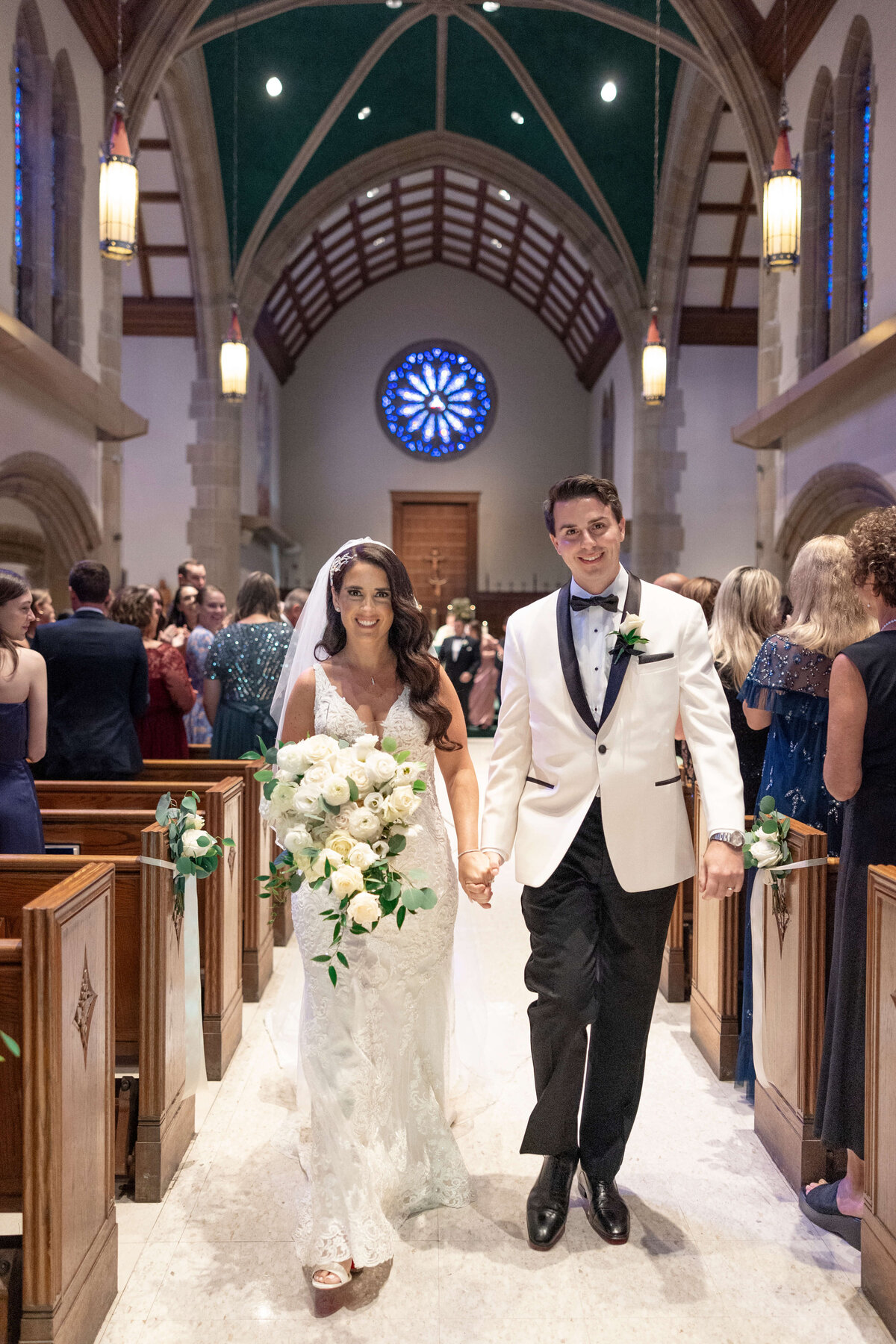 A bride in a white lace gown and veil holds a bouquet of white flowers, walking hand in hand with a groom in a white tuxedo with a black bow tie. They are in a church with high ceilings and a stained glass window, surrounded by seated guests.