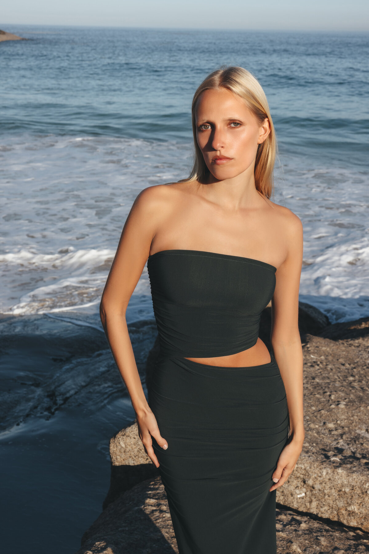 Model Johanna staat in een zwarte cut-out jurk op een rots aan zee in Kaapstad, met haar blik gericht op de camera en de golven die zachtjes achter haar breken.